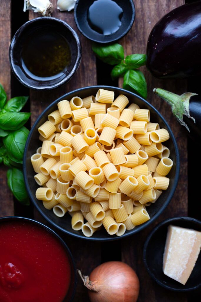 Zutaten für Pasta alla Norma in Schüsseln auf dunkelm Holzuntergrund. Zu sehen sind Rigatoni, gehackte Tomaten, Basilikum, ein Stück Parmesan, Aubergine, Knoblauchzehen, eine Zwiebel, Olivenöl und Ahornsirup.