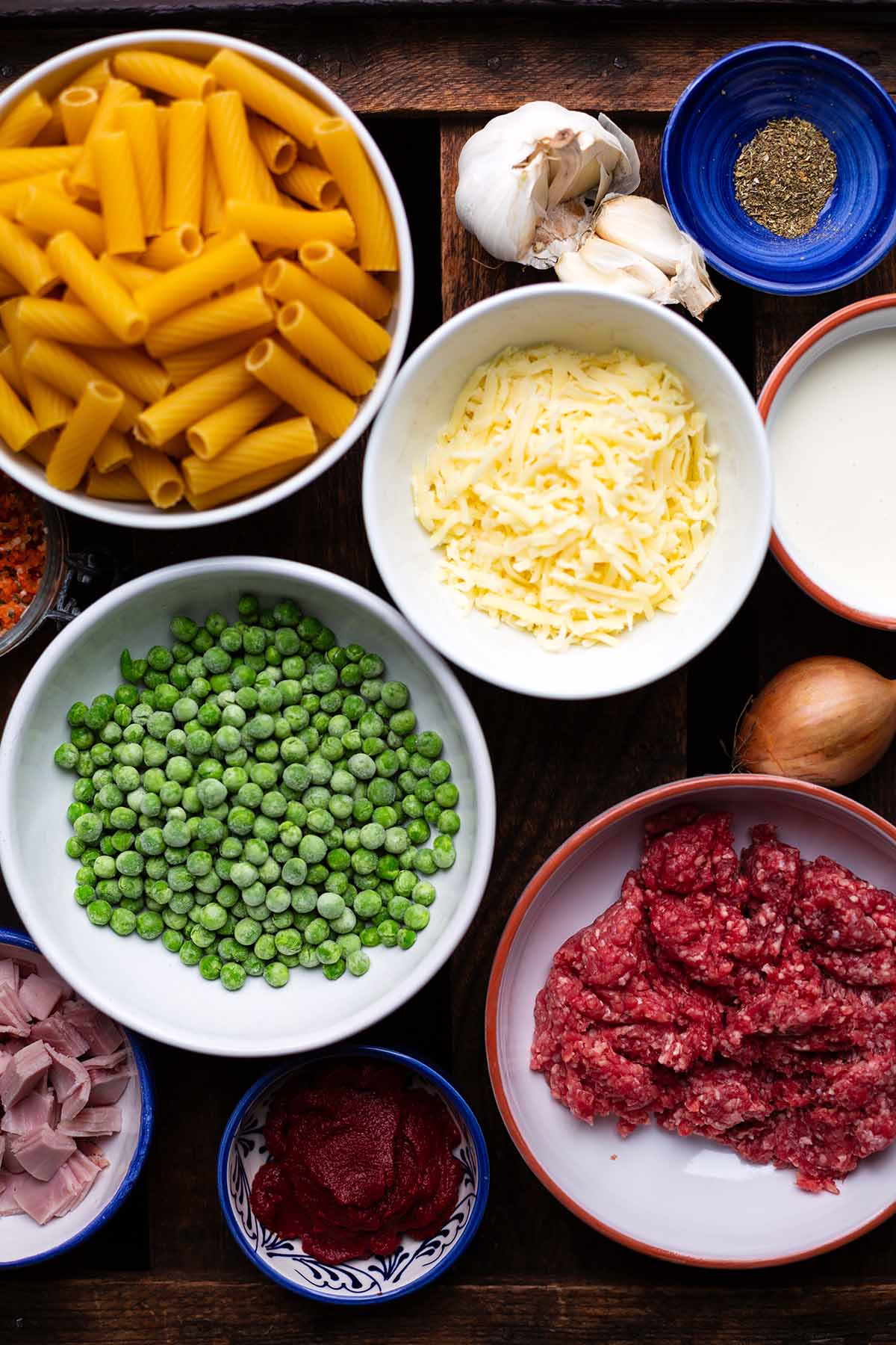 Die Zutaten für italienischen Nudelauflauf Rigatoni al Forno: Rigatoni, Gewürze, Käse, Erbsen, Hackfleisch, Tomatenmark und Schinken.