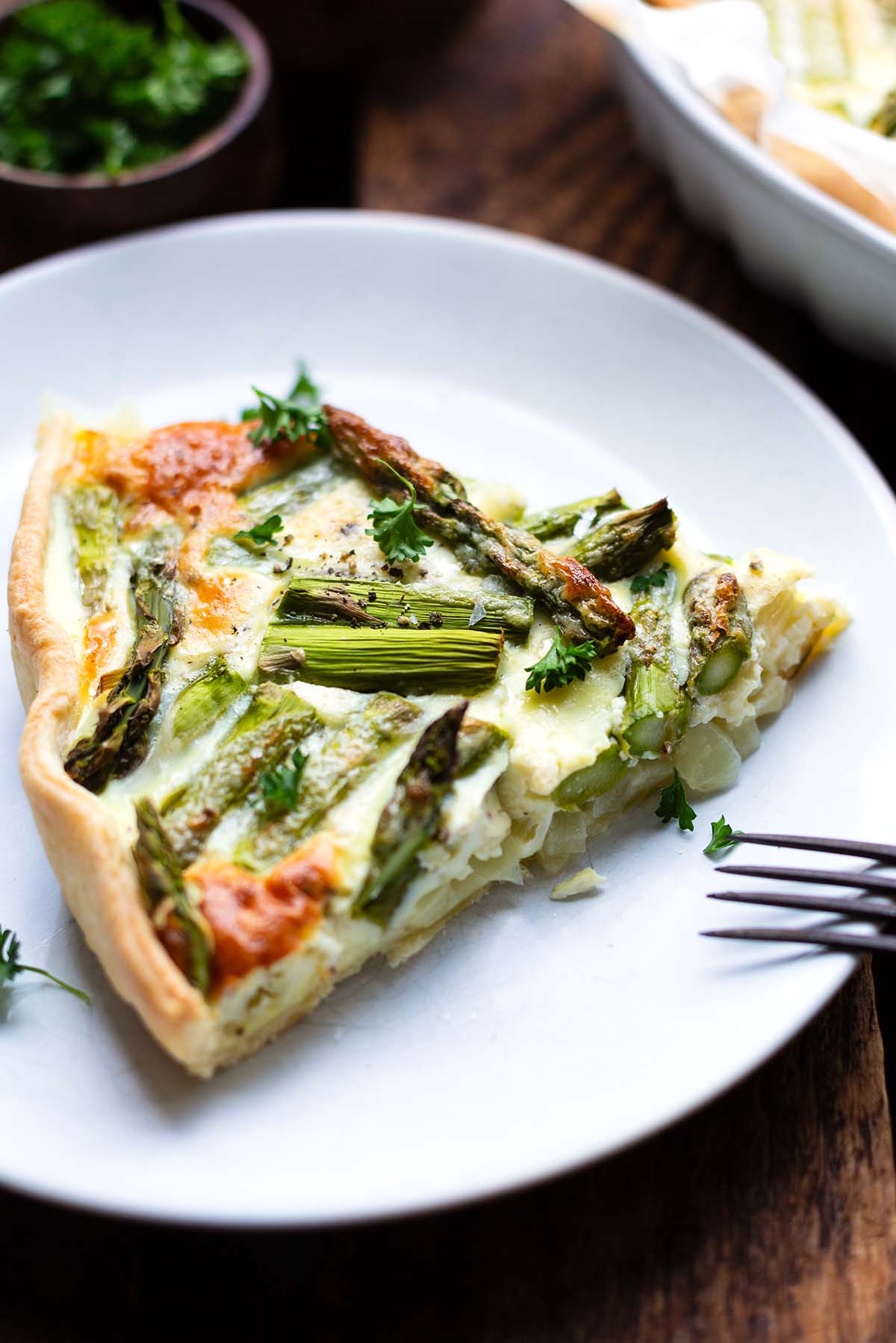 Ein dreieckiges Stück der fertigen Spargel-Quiche auf einem weißen Teller. Garniert mit Petersilie.