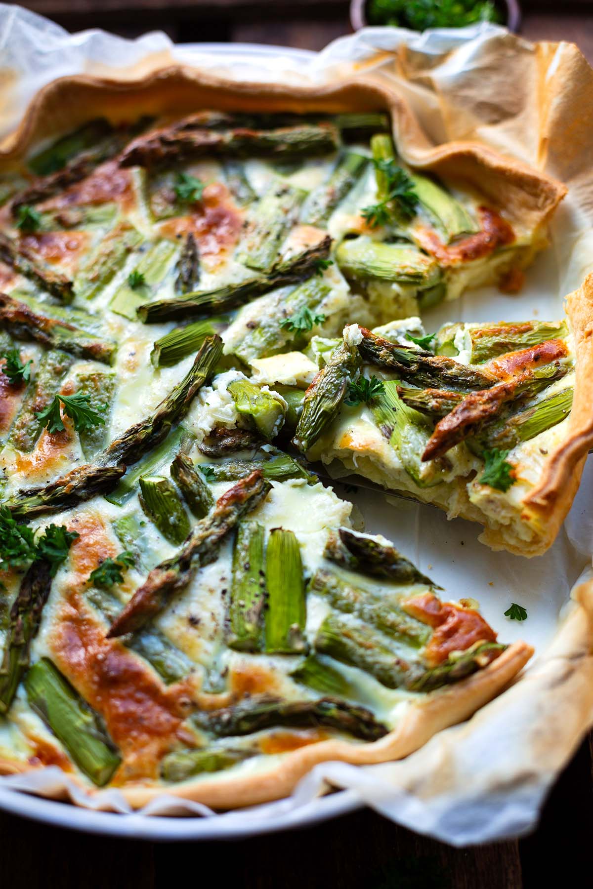 Fertige Spargel-Quiche mit grünem Spargel in weißer Aufflaufform auf dunklem Holzuntergrund. Im Hintergrund eine kleine braune Schüssel mit Petersilie. Ein dreieckiges Stück wird mit einem Tortenheber angehoben.