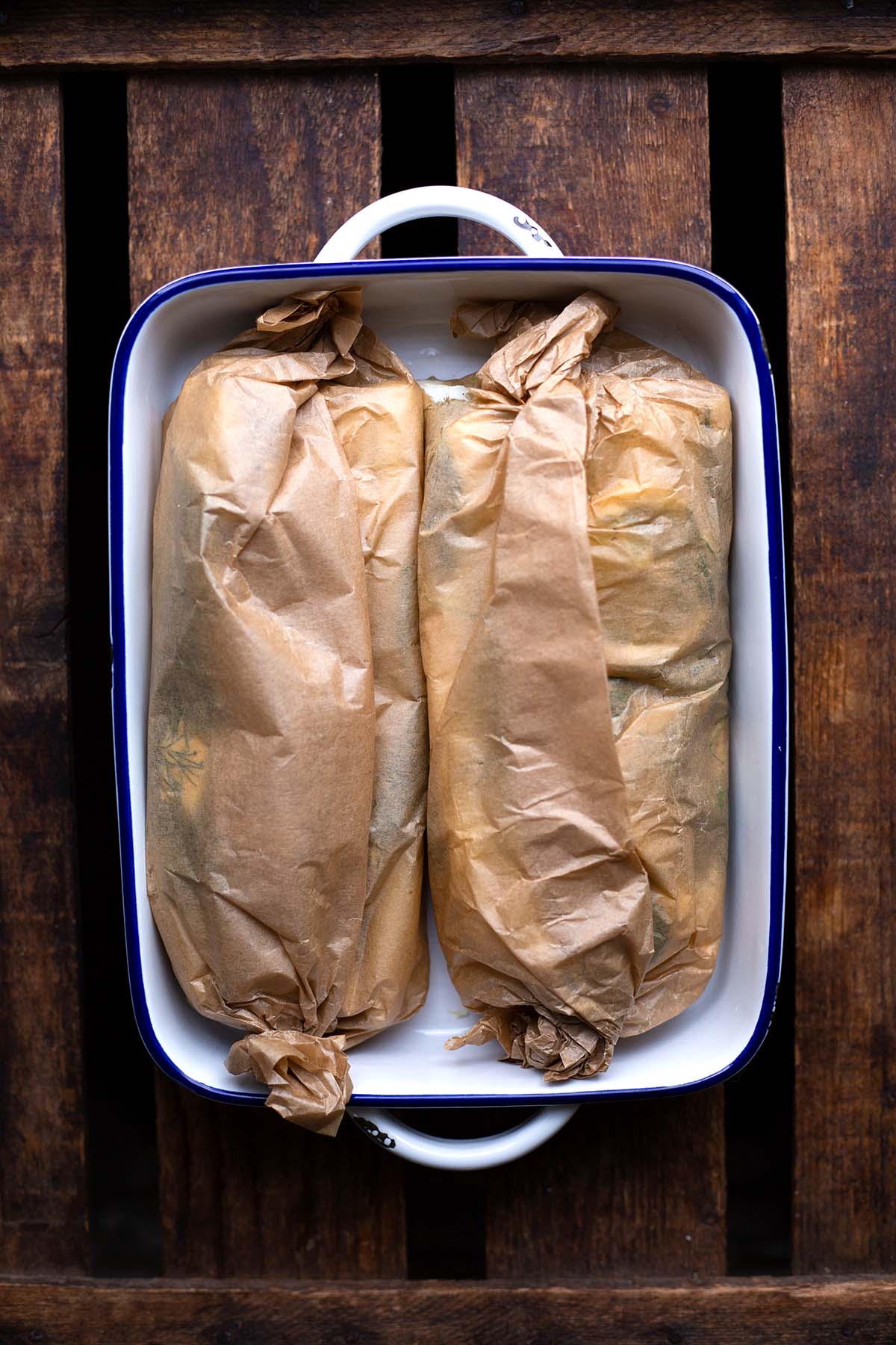 Spargel eingepackt in zwei Backpapier-Päckchen in einer weißen Auflaufform mit blauem Rand auf dunklem Holzuntergrund.