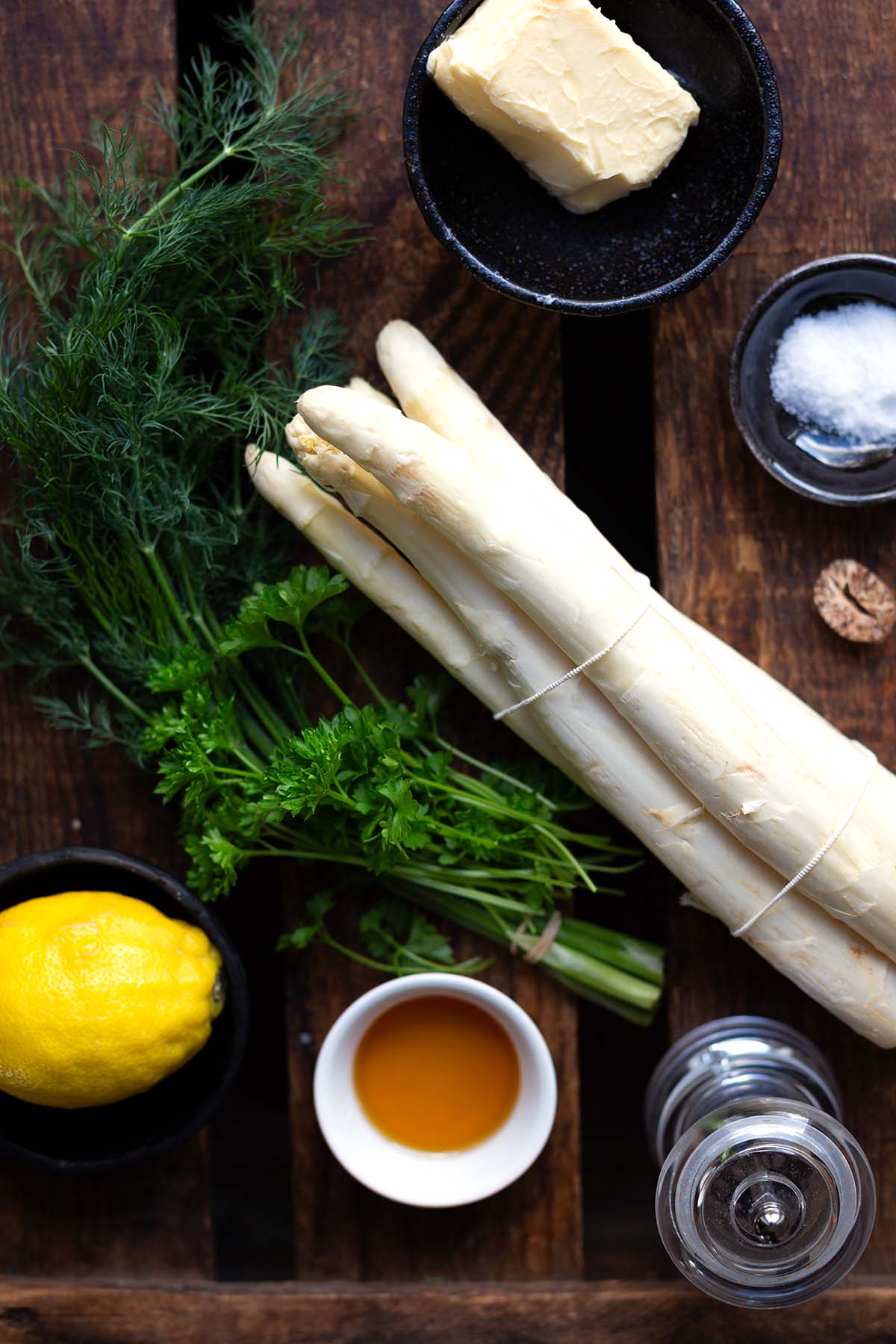 Die Zutaten für den weißen Spargel aus dem Ofen auf einem dunkeln Holzuntergrund. Zu sehen sind: eine Zitrone, Dill, Petersilie, ein Bund weißer Spargel, Ahornsirup, Butter, Salz jeweils in einer weißen Schüssel, eine Pfeffermühle und Muskat.