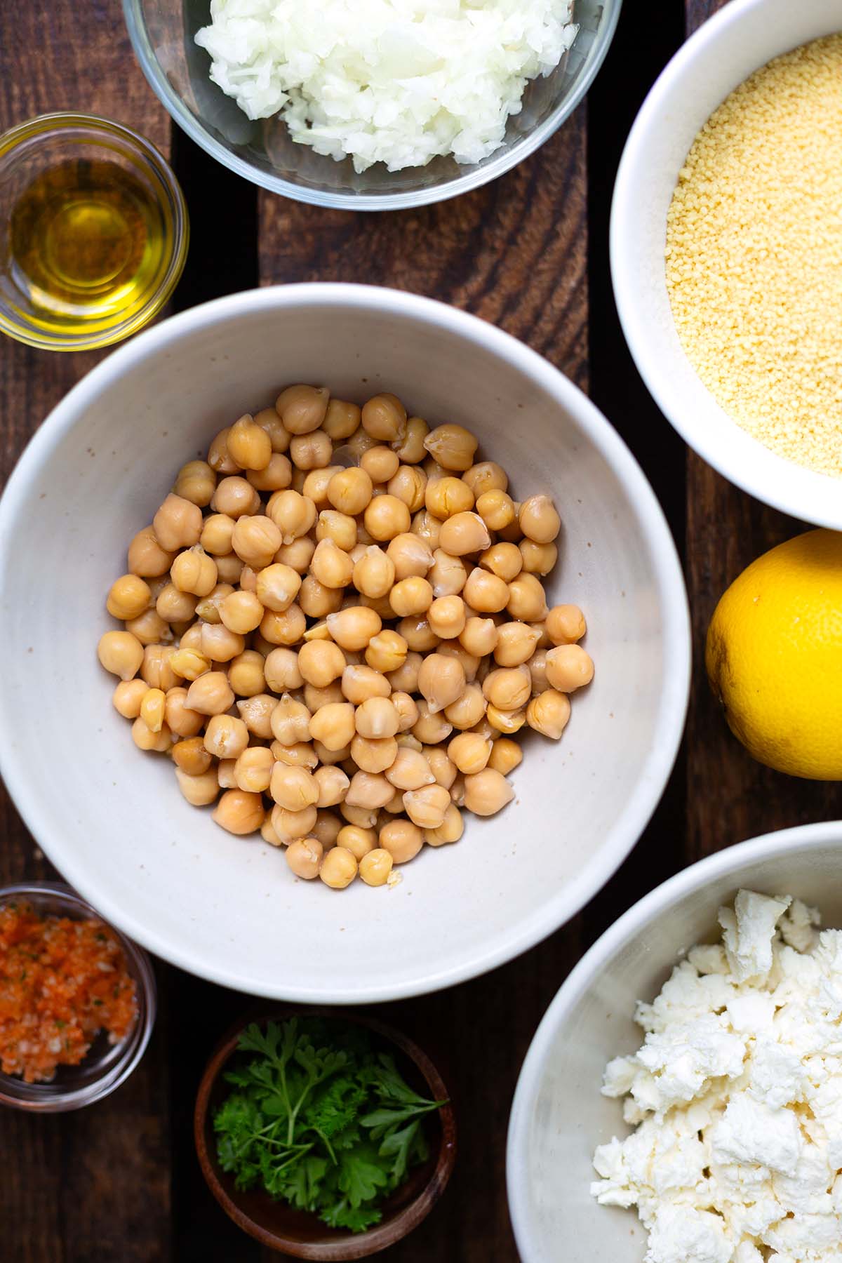 DIe Zutaten für den Couscous-Salat verteilt in Schüsseln auf dunklem Holzuntergrund. ZU sehen sind, Kichererbsen, Gemüsebrühe, zerbröselter Feta, Zitrone, Couscous, Zwiebeln und Öl.