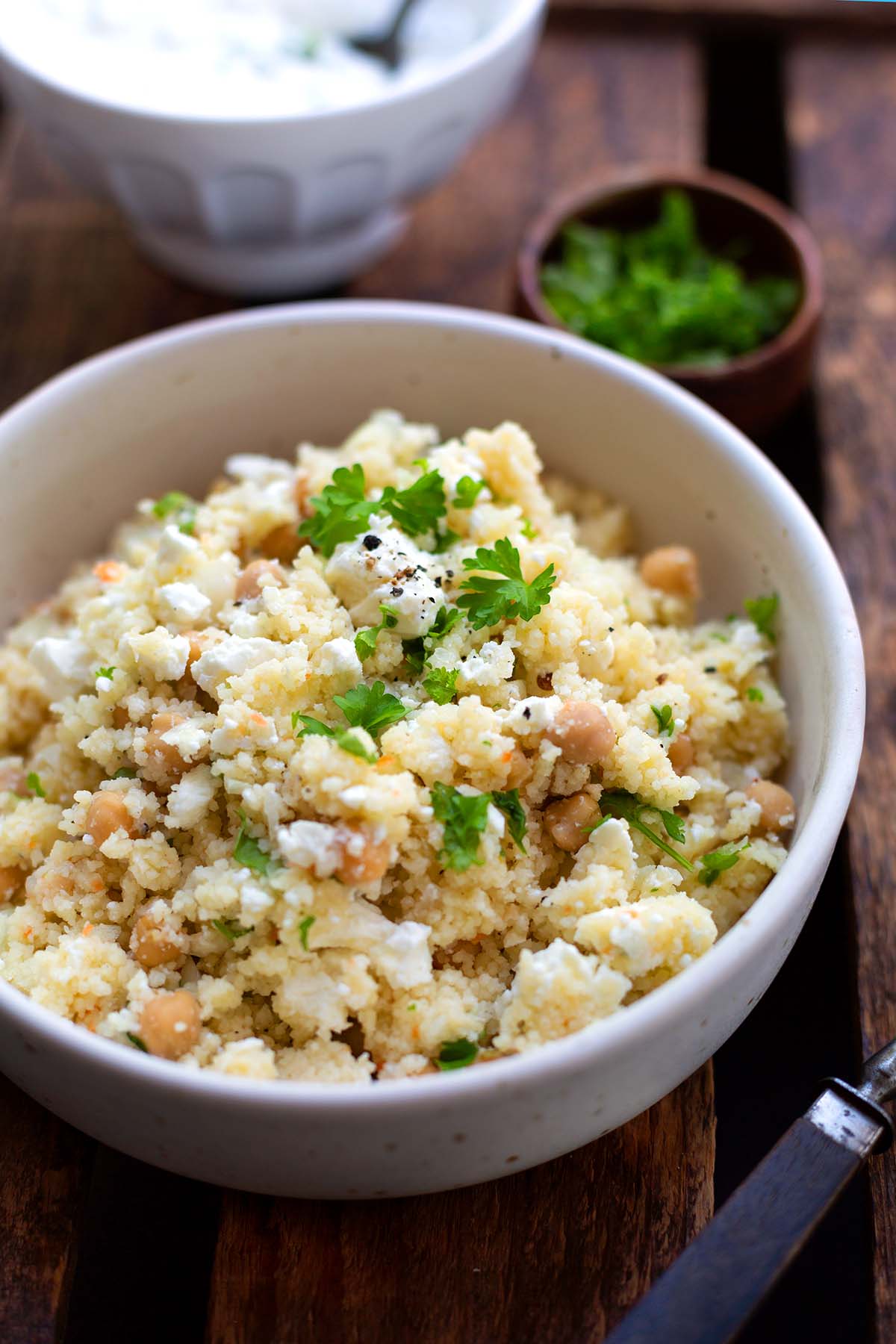 Der fertige Couscous-Salat mit Feta in einer weißen Schüssel auf dunklem Holzuntergrund und garniert mit Petersilie und einer Prise Pfeffer.