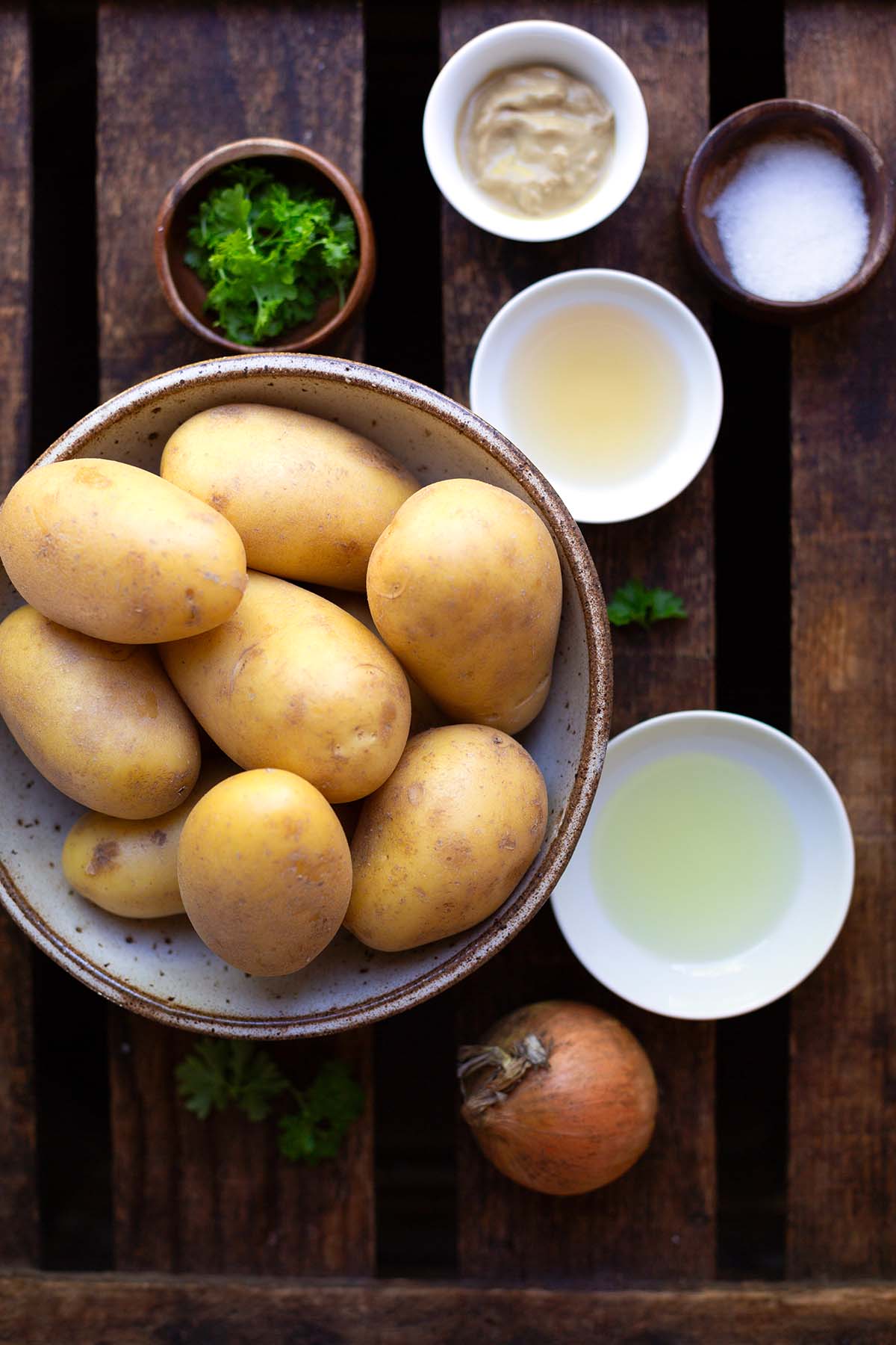 Zutaten für den Kartoffelsalat ohne Mayo in Schüsseln auf dunklem Holzuntergrund. Zu sehen sind rohe, ungeschälte Kartoffeln, Petersilie, Zucker, Öl, Weißweinessig, weiße Zwiebel, Senf.