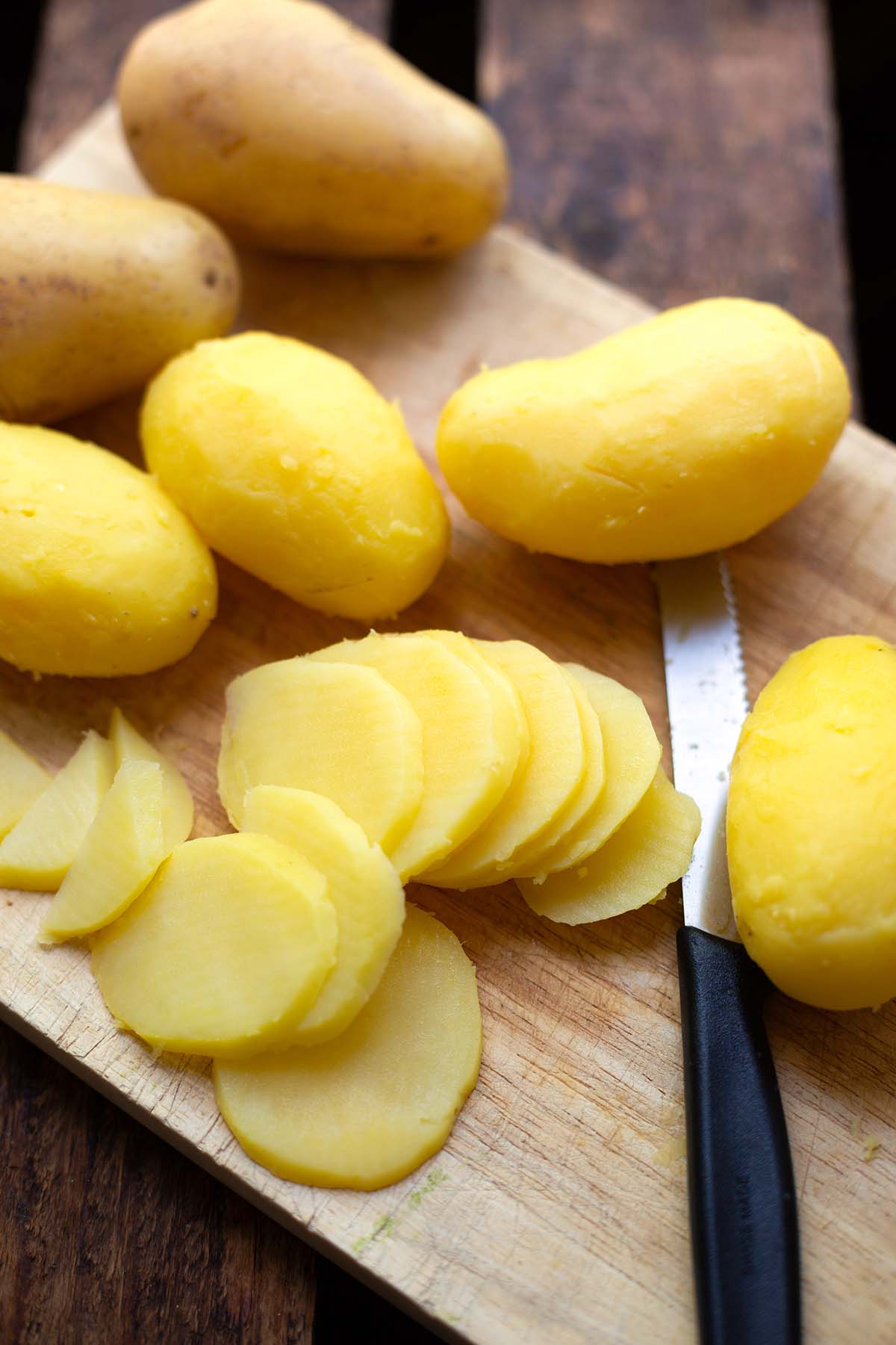 Pellkartoffeln auf einem Holzbrett. Zwei sind im Hintergrund noch ungeschält. Die anderen Kartoffeln für den Kartoffelsalat sind gepellt und eine bereits in Scheiben geschnitten. Das Messer liegt noch daneben.