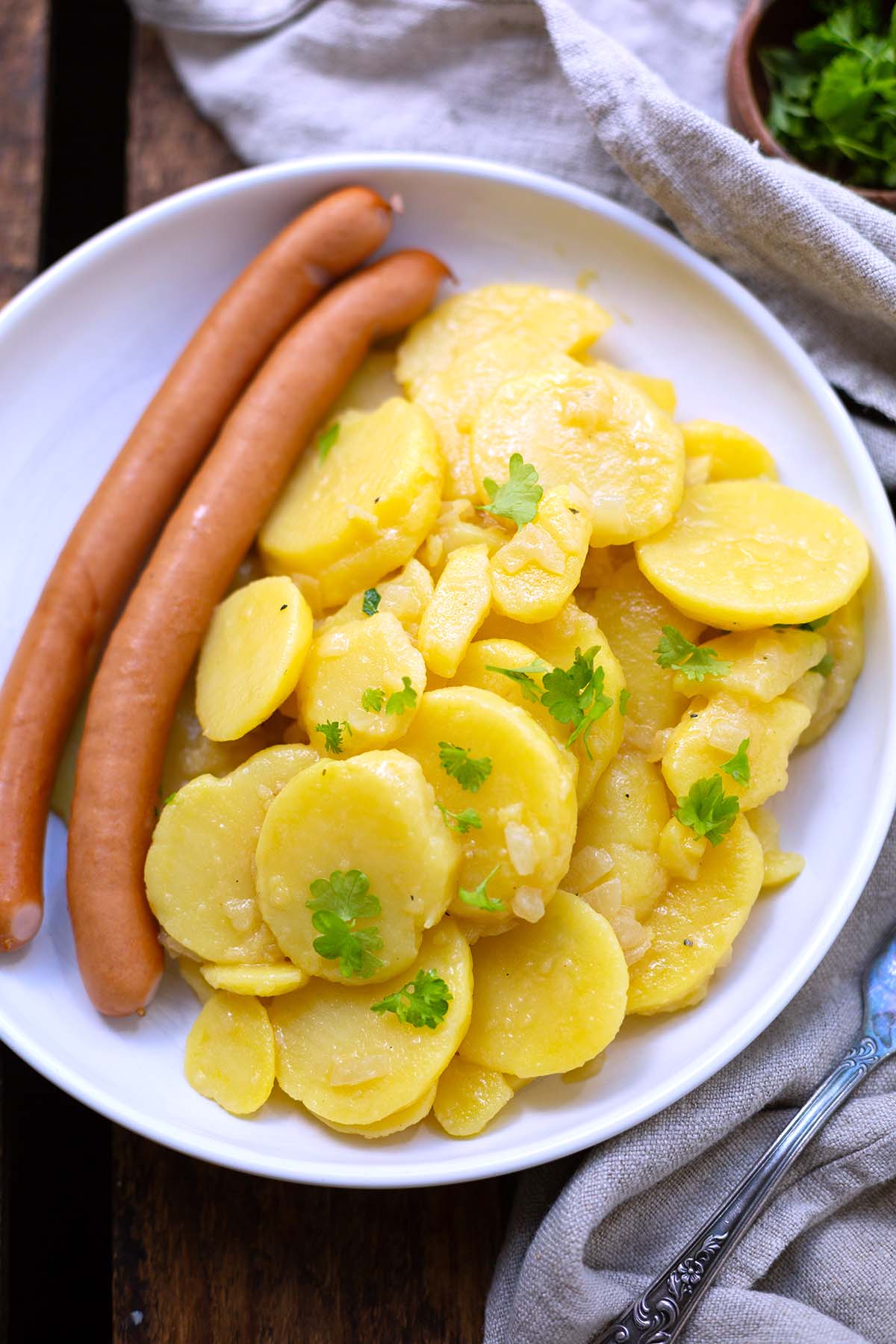 Fertiger Kartoffelsalat ohne Mayo angerichtet auf einem weißen Teller mit zwei Wiener Würstchen und garniert mit Petersilie.