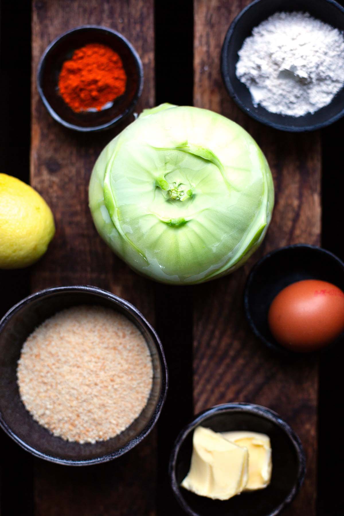 Zutaten für einfache Kohlrabischnitzel auf einem dunkeln Holzuntergrund aufgeteilt auf einzelne dunkle Schüsseln.
Zu sehen sind ein großer Kohlrabi, Paprikapulver, Mehl, Zitrone, Paniermehl, ein Ei und Butter.
