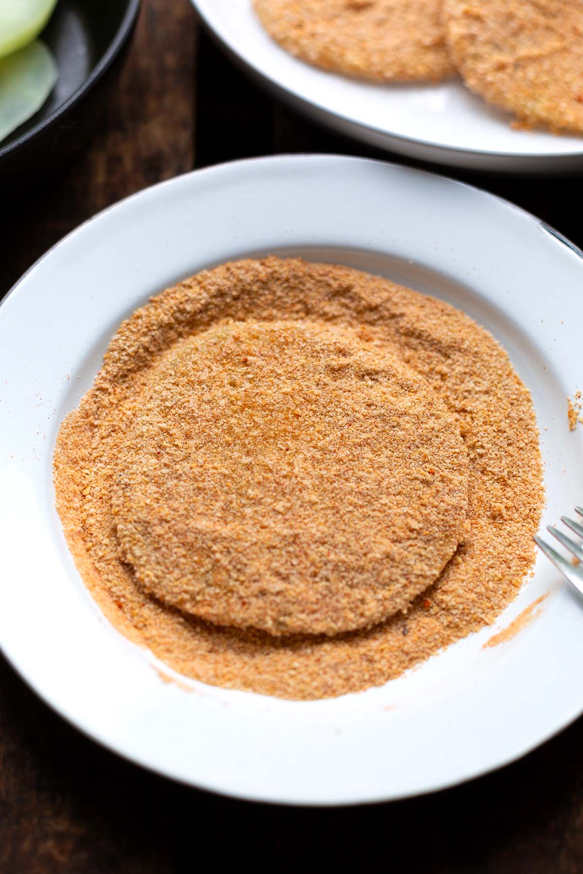 Paniermehl auf einem weißen Teller. Darin liegt eine schon panierte Kohlrabischeibe.