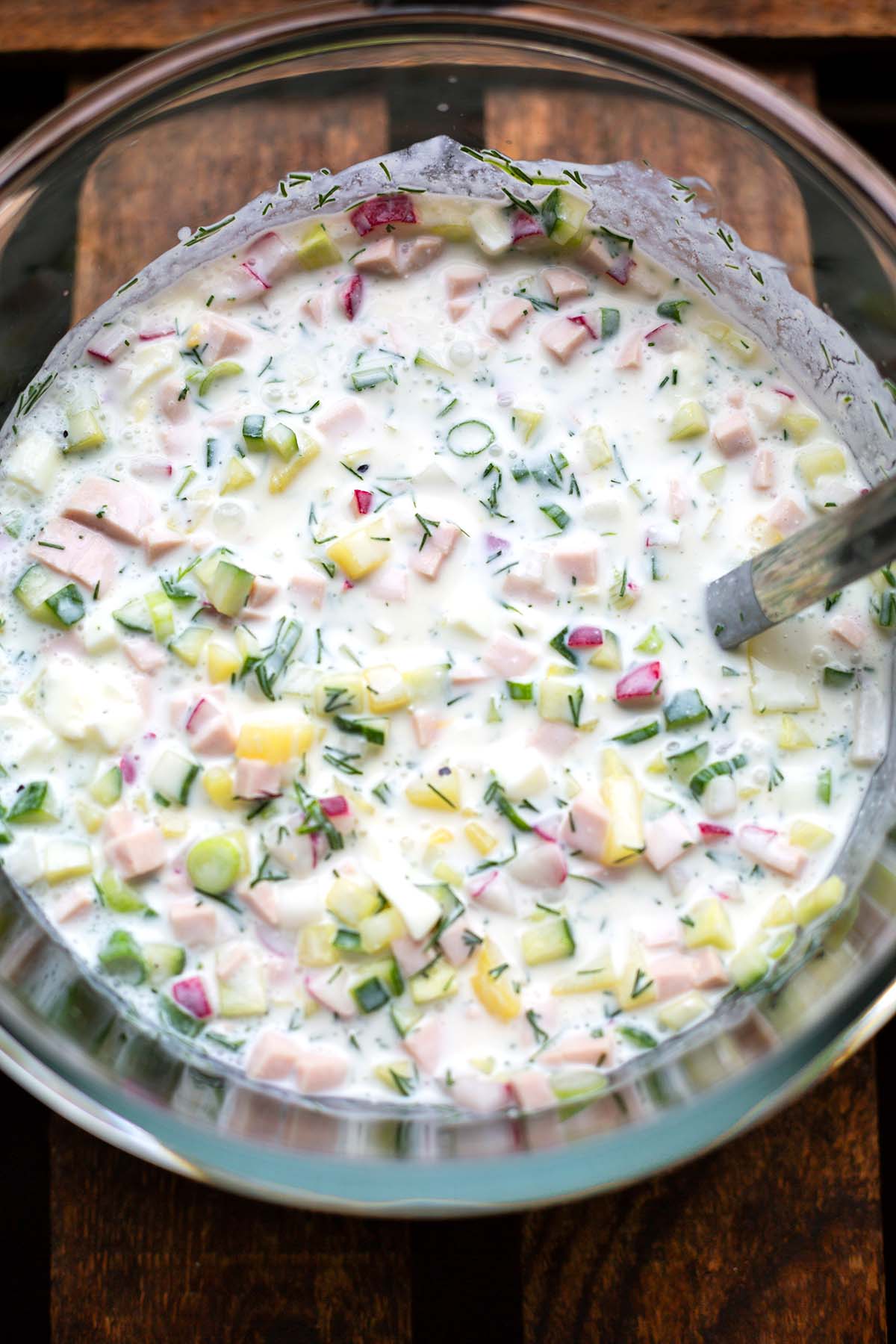 Okroschka in einer Glasschüssel mit Fleischwurst, Kartoffeln, Ei und Radieschen mit einer Suppenkelle.