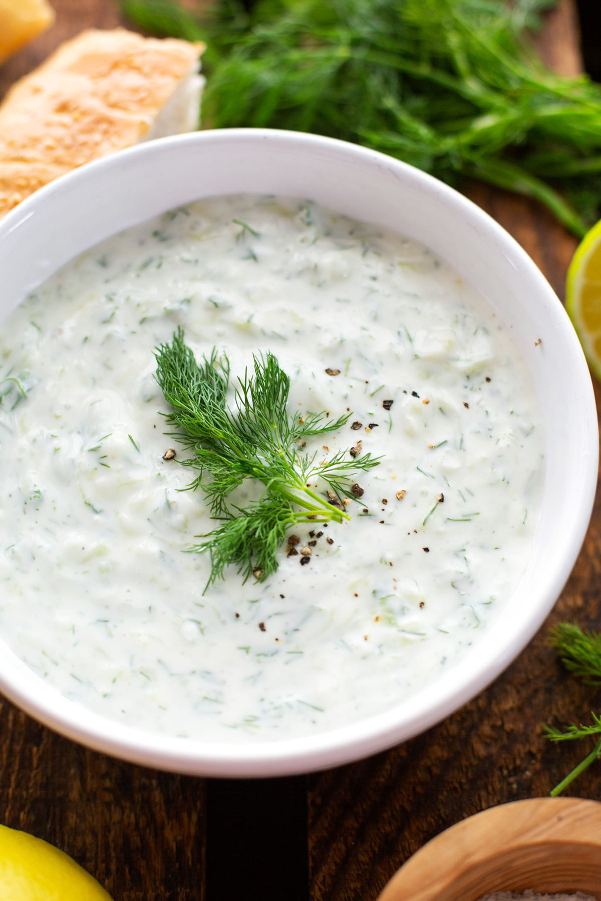 Tzatziki in einer weißen Schüssel. Getoppt mit Dill, im Hintergrund Fladenbrot und Zitrone