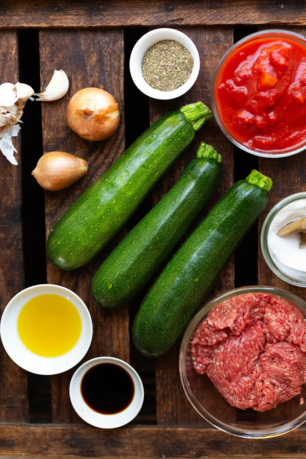 Zutaten für die Zucchini-Hackfleisch-Pfanne in einzelnen Schälchen oder direkt auf einem dunklen Holzuntergrund. Zu sehen sind drei Zucchini, gehackte Tomaten, Hackfleisch, Zwiebel, Knoblauch, Ö, Sojasauce und Gewürze.