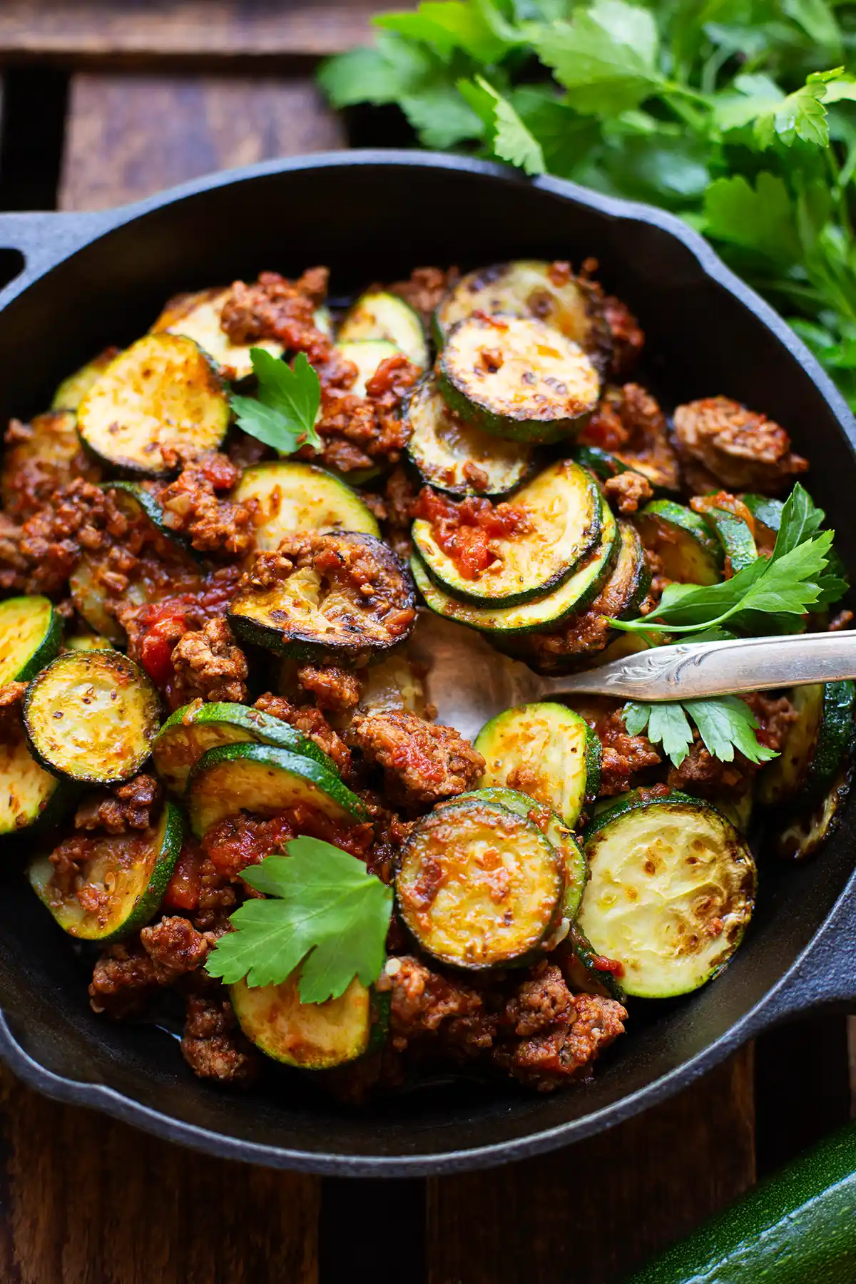 Fertige Zucchini-Hackfleisch-Pfanne in einer schwarzen Pfanne auf dunklem Holzuntergrund. Garniert mit glatter Petersilie. Ein Bund frische Petersilie liegt neben der Pfanne. Eine Gabel steckt in der low carb Zucchini-Pfanne.