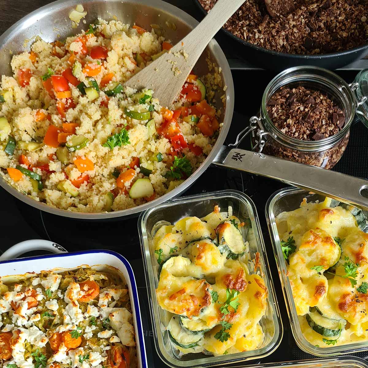 Blick auf viele verschiedene vorgekochte Gerichte. Zwei Dosen mit Kartoffelauflauf mit Zucchini und Käse überbacken, Couscouspfanne mit Gemüse, Granola mit Schokolade und ein weiterer Auflauf.