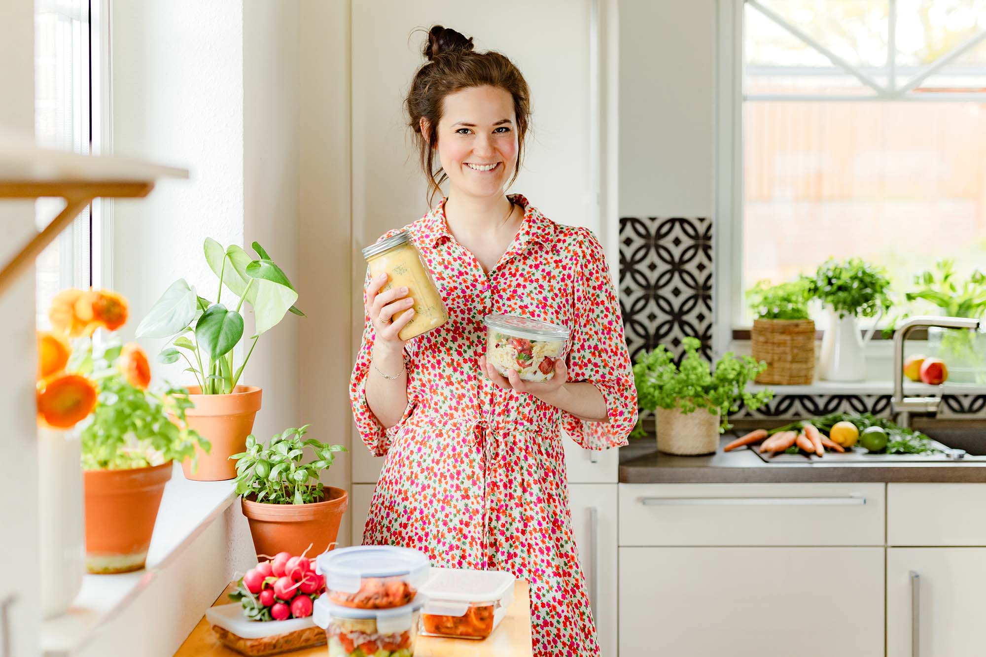 Mia in einem roten Blumenkleid steht in einer weißen Küche. In jeder Hand hält sie vorgekochtes Essen in einer Dose. Auf dem Tisch vor ihr stehen weitere Dosen mit vorgekochtem Essen. Im Hintergrund auf der Spüle liegt verschiedenes Gemüse.