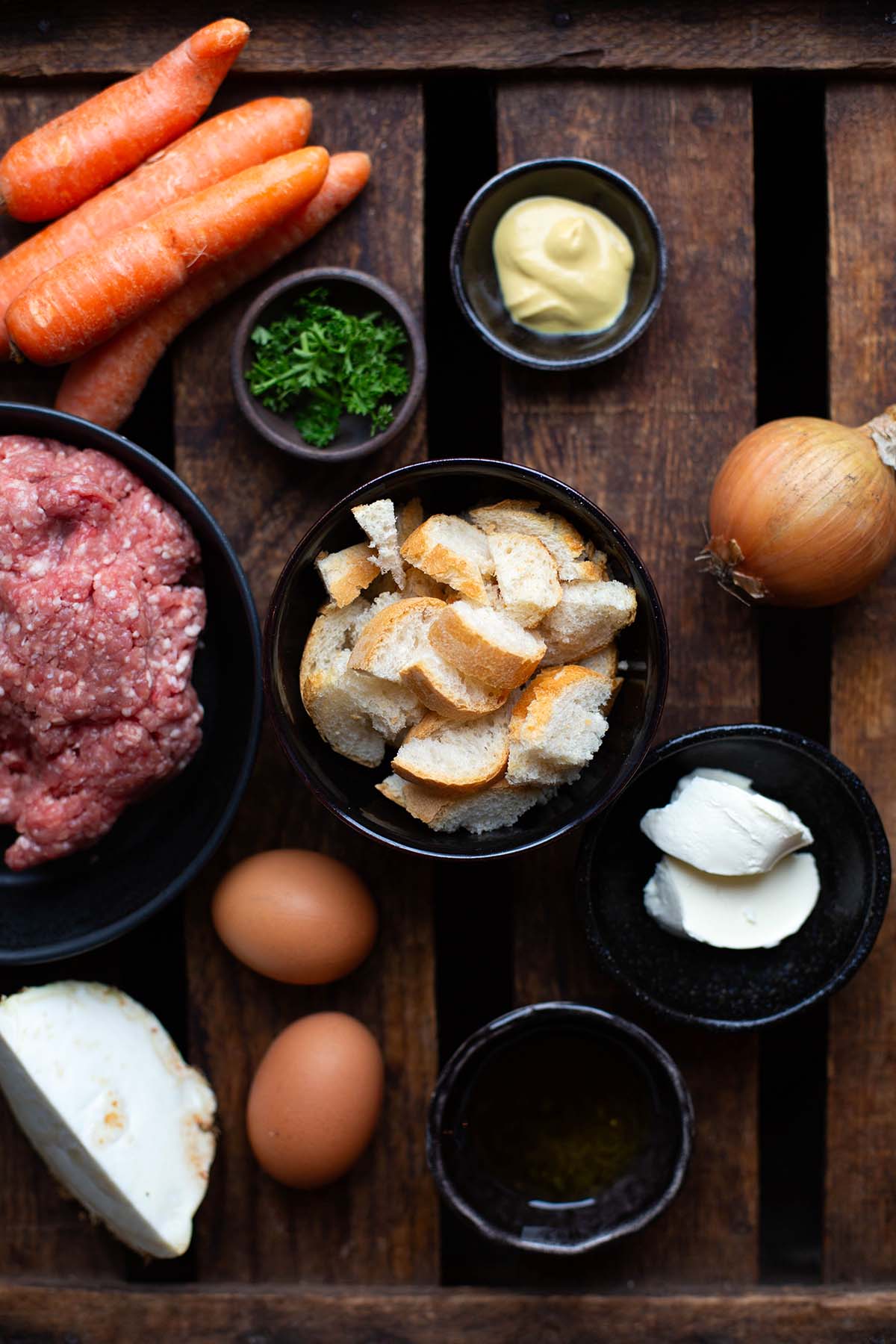 Die Zutaten für Hackbraten mit Soße auf einer braunen Holzkiste: Hackfleisch, Karotten, Zwiebel, Brötchen, Petersilie, Sellerie, Öl, Eier, Creme Fraiche