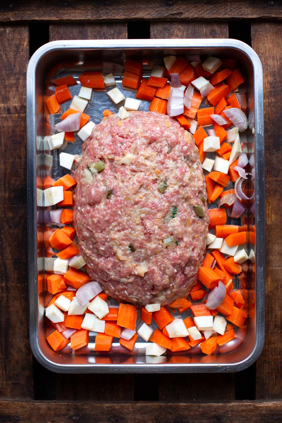 Hackbraten vor dem Backen in der Auflaufform. Drumherum kleingewürfeltes Gemüse für die Soße