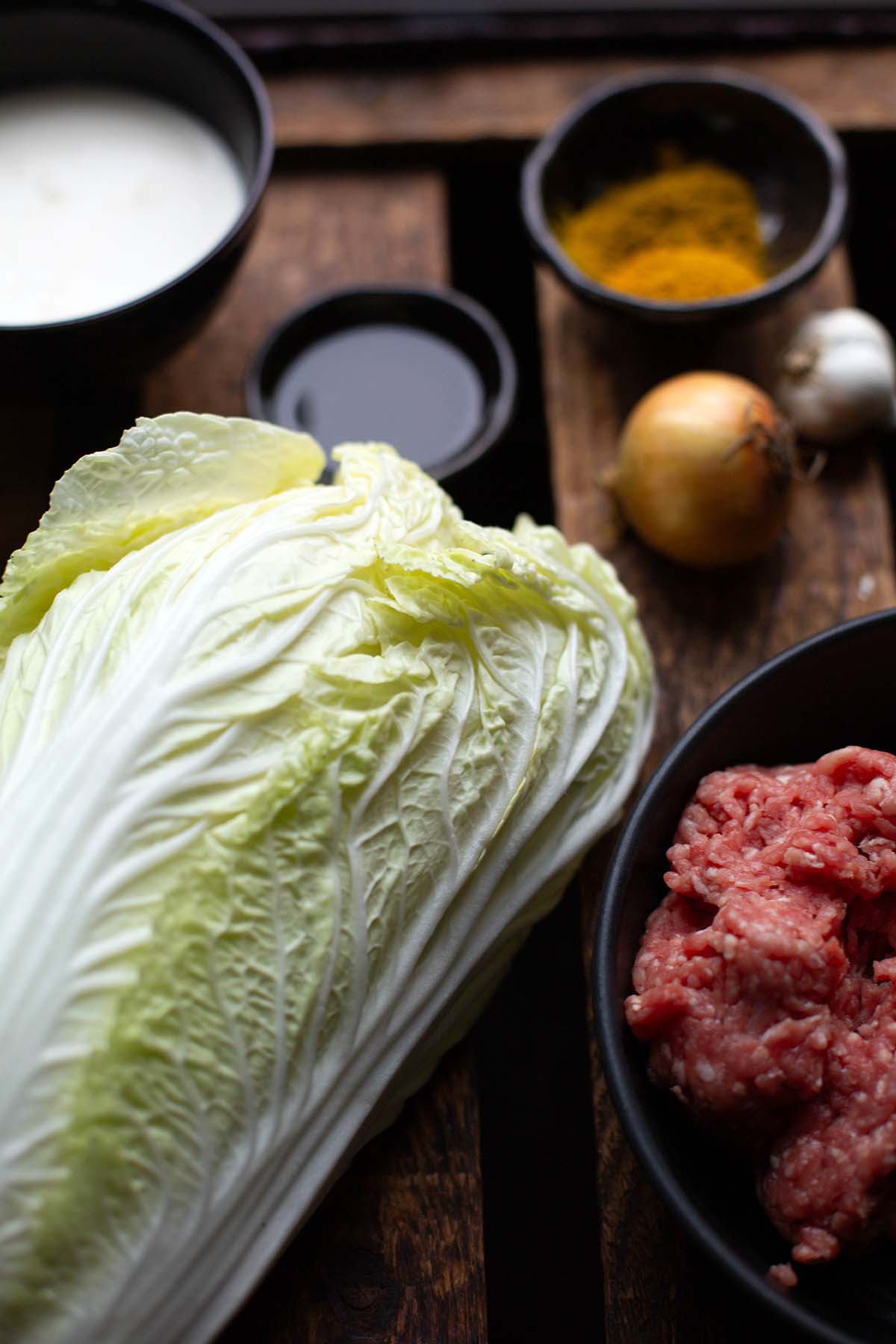 Zutaten für die Chinakohl-Pfanne auf dunklem Holzuntergrund. Zu sehen sind Chinakohl, Zwiebel, Knoblauch, Hackfleisch, Currypulver, Sahne und Olivenöl.