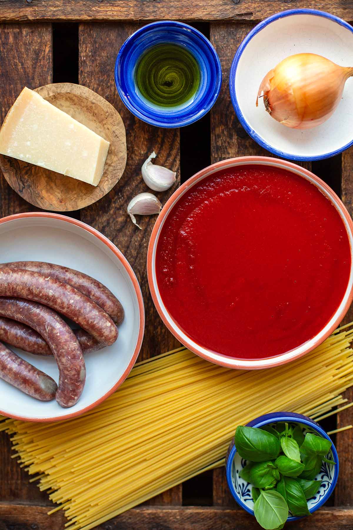 Zutaten für die Spaghetti mit Fleischbällchen aufgeteilt auf Schüsseln, die auf dunklem Holzuntergrund stehen. Zu sehen sind passierte Tomaten, Zwiebel, Parmesan, Olivenöl, grobe Bratwürstchen, Knoblauchzehen, Spaghetti und Basilikum.