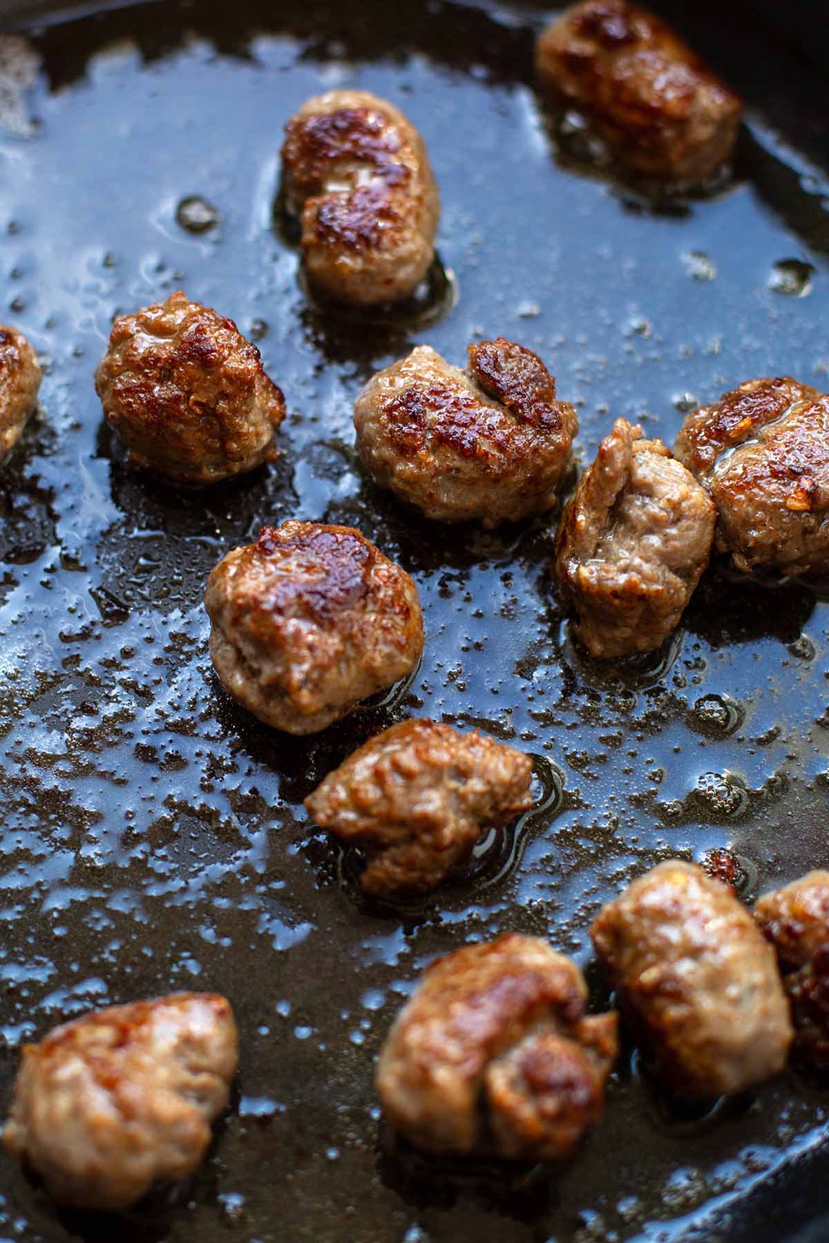 Zu Kugeln gerolltes Brät aus groben Bratwürstchen werden in einer schwarzen Pfanne mit Öl gebraten.