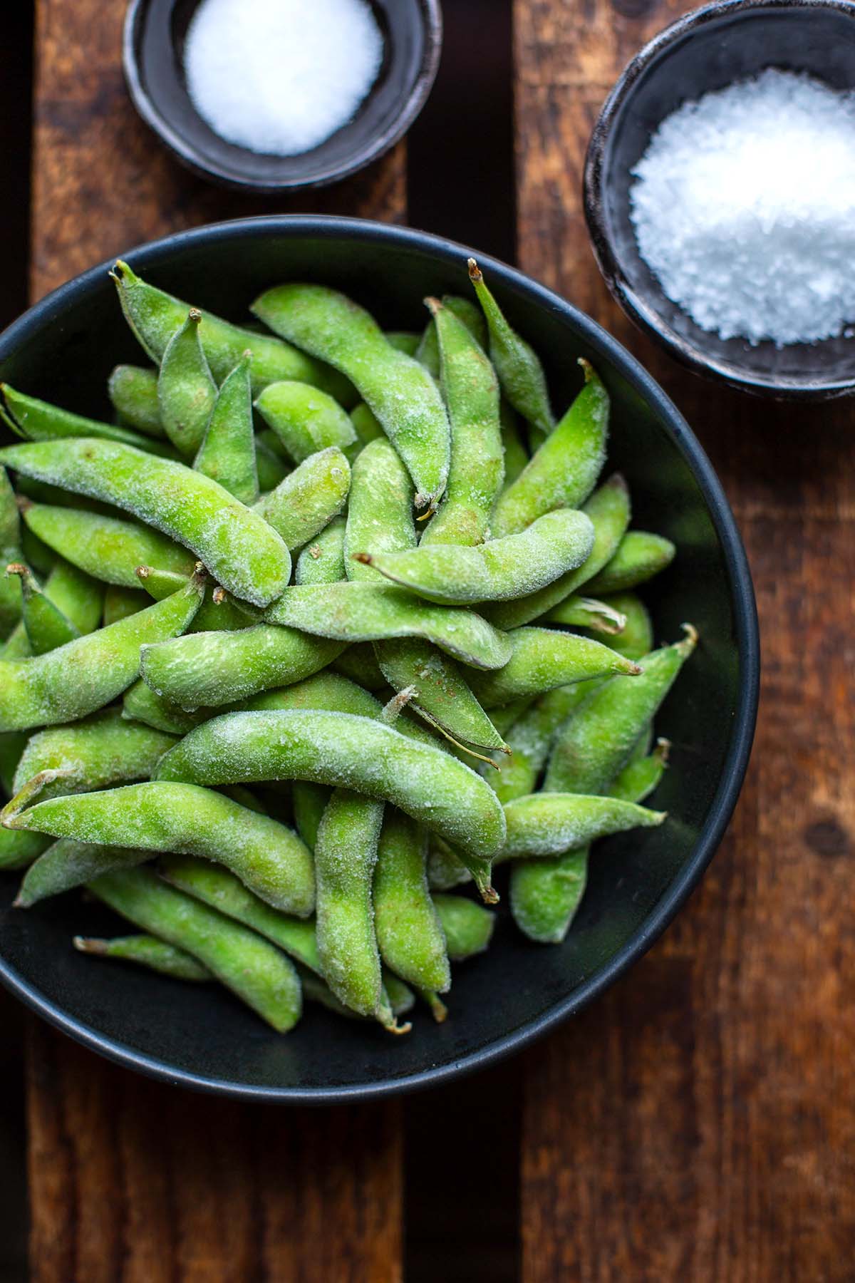 Die Zutaten zum Edamame zubereiten: TK-Edamame mit Schale, Salz und Meersalz auf dunklem Holzuntergrund.
