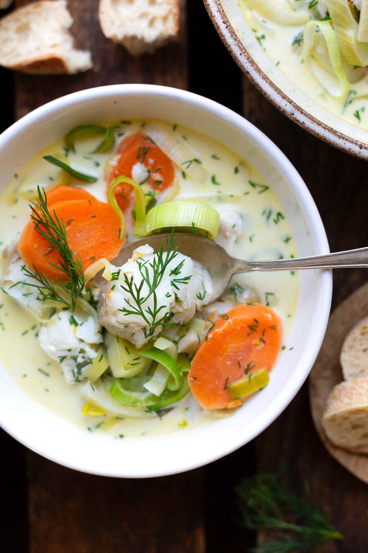 Fischsuppe mit Gemüse, Zitrone und Dill in einer weißen Schüssel auf dunklem Untergrund. Dazu Baguette.