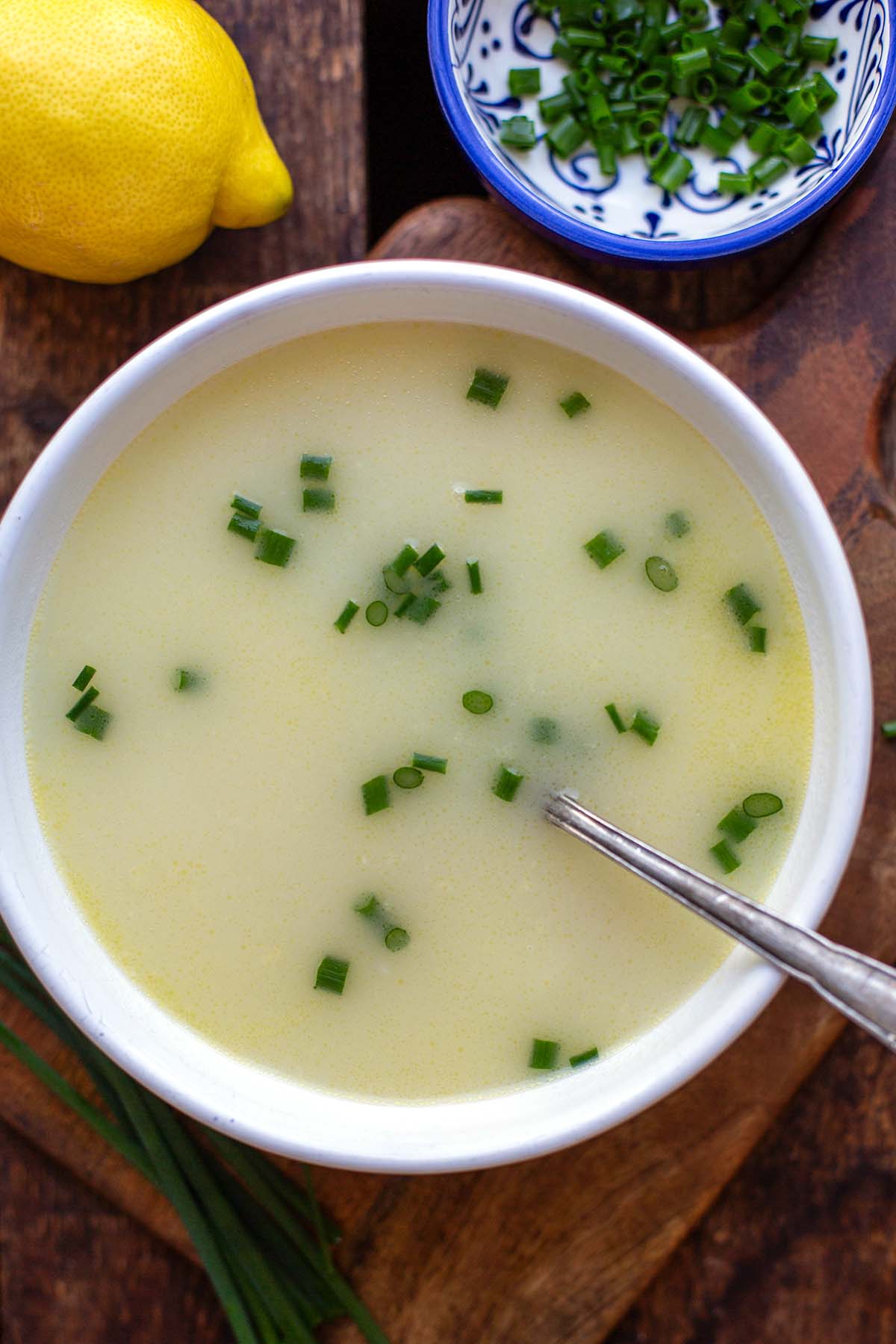 Fertige Spargelsuppe aus Schalen in einer weißen Schüssel auf dunklem Holzuntergund und mit einem silbernen Löffel. Garniert mit Schnittlauch. Im Hintergrund ist eine ganze Zitrone und noch eine blau-weiße Schale mit geschnittenem Schnittlauch.