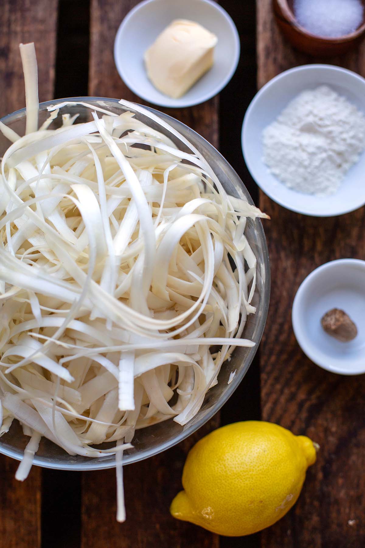 Einzelne Zutaten für die Spargelsuppe aus Schalen auf einem dunklen Holzuntergrund. Zu sehen sind Spargelschalen, Zitrone, Mehl, Butter, Zucker und Muskatnuss.