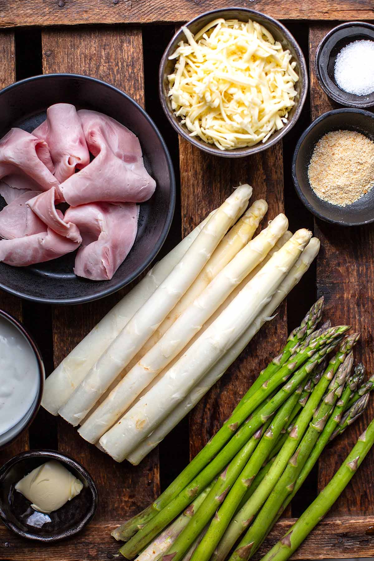 Die einzelnen Zutaten für überbackenen Spargel. Zu sehen sind weißer und grüner Spargel, Schinken, Semmelbrösel, geriebener Käse, Salz, Butter und Béchamelsoße.