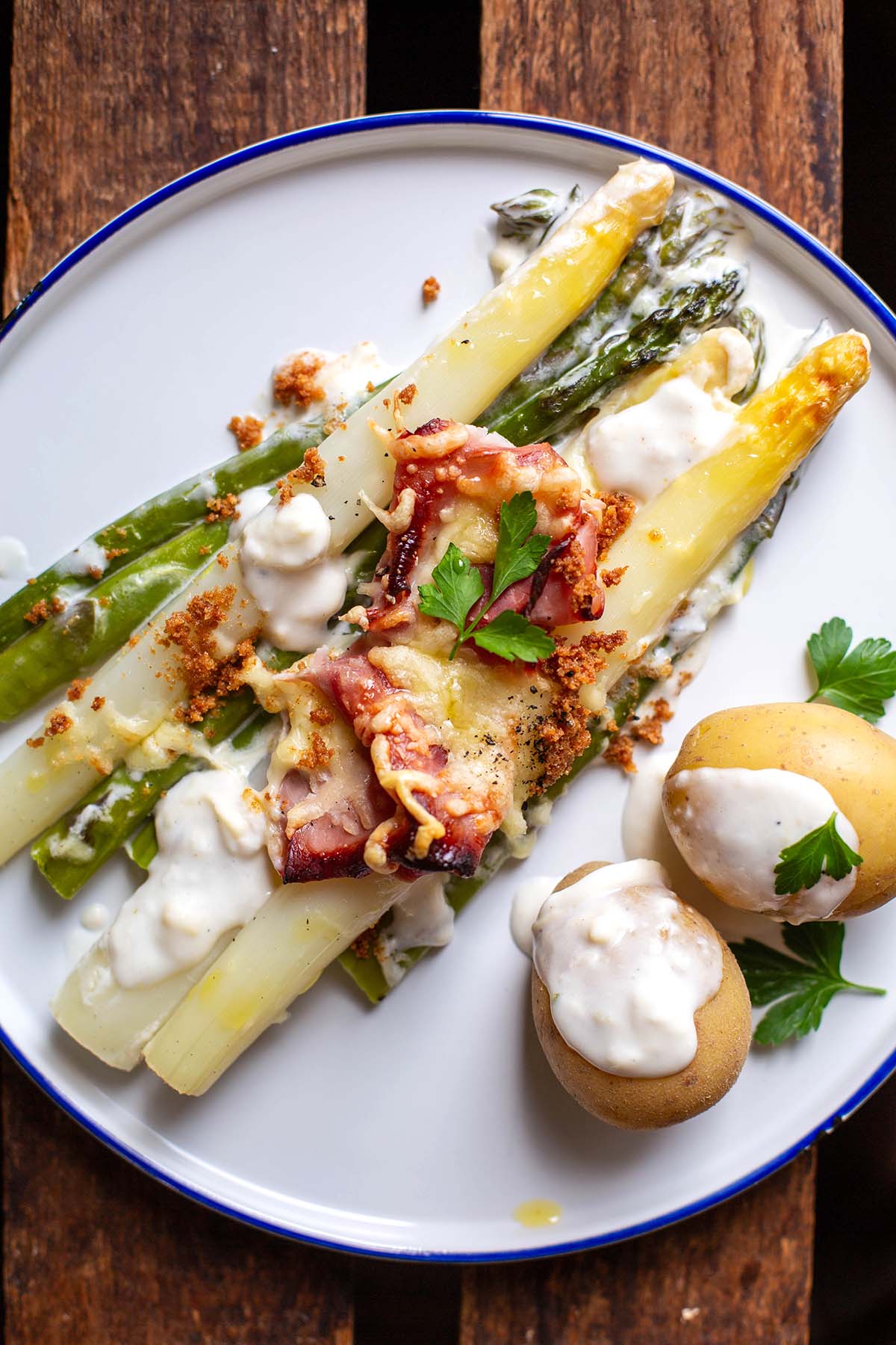 Überbackener Spargel (grüner und weißer Spargel auf einem weißen Teller mit einem blauen Ran. Auf dem Teller ebenfalls zwei Pellkartoffeln mit Soße und garniert mit Petersilie.