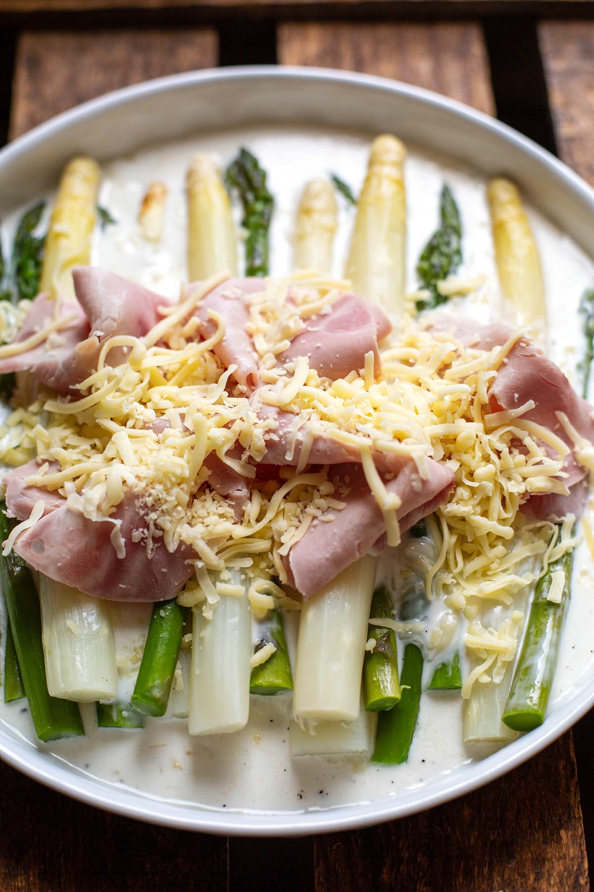 Grüner und weißer Spargel auf einem weißen Teller zusammen mit Schinken, Käse und Béchamelsoße bevor das Ganze in den Backofen kommt.