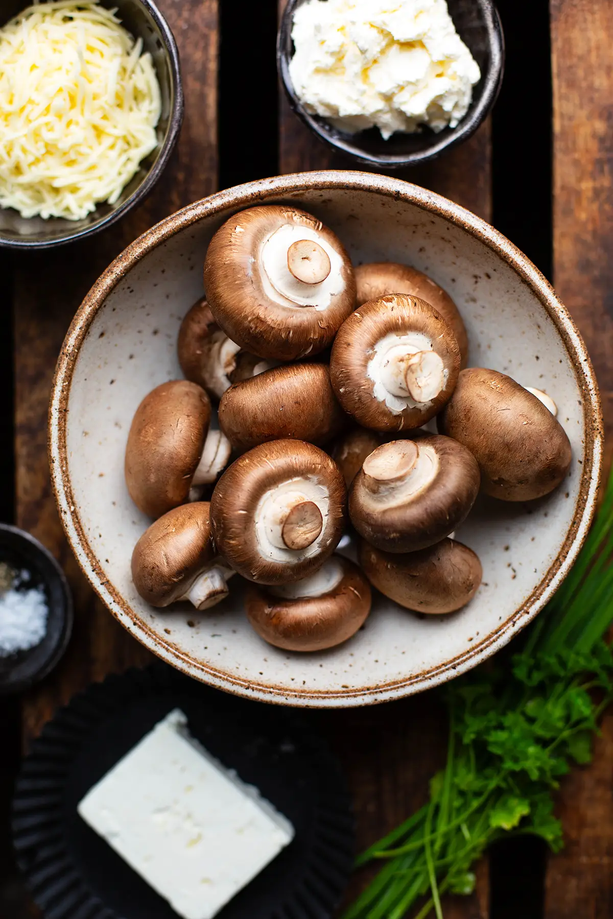 Zutaten für gefüllte Champignons. Zu sehen sind braune Champignons, Salz, Feta, geriebener Käse, Schnittlauch und Petersilie. Die Zutaten stehen in kleinen Schüsseln auf einem dunklen Holzuntergrund.