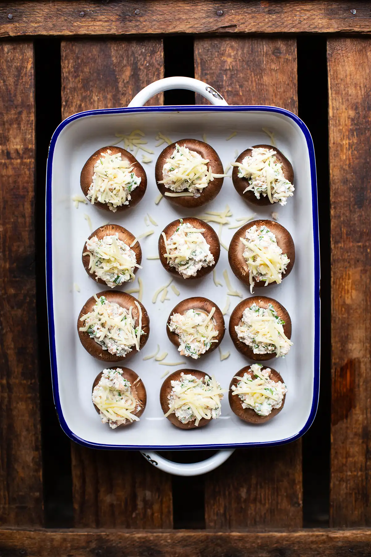 12 vorbereitete Champignons mit vegetarischer Füllung in einer weißen Auflaufform mit blauem Rand. Die Auflaufform steht auf einem dunklen Holzuntergrund.