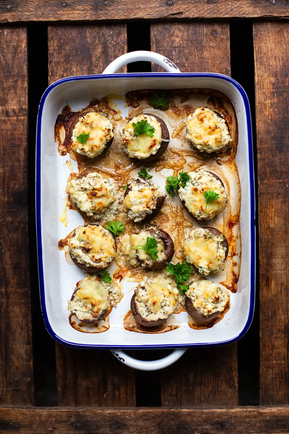 Die fertig gebackenen vegetarischen Champignons in einer weißen Auflaufform mit blauem Rand. Die Auflaufform steht auf einem dunklen Holzuntergrund.