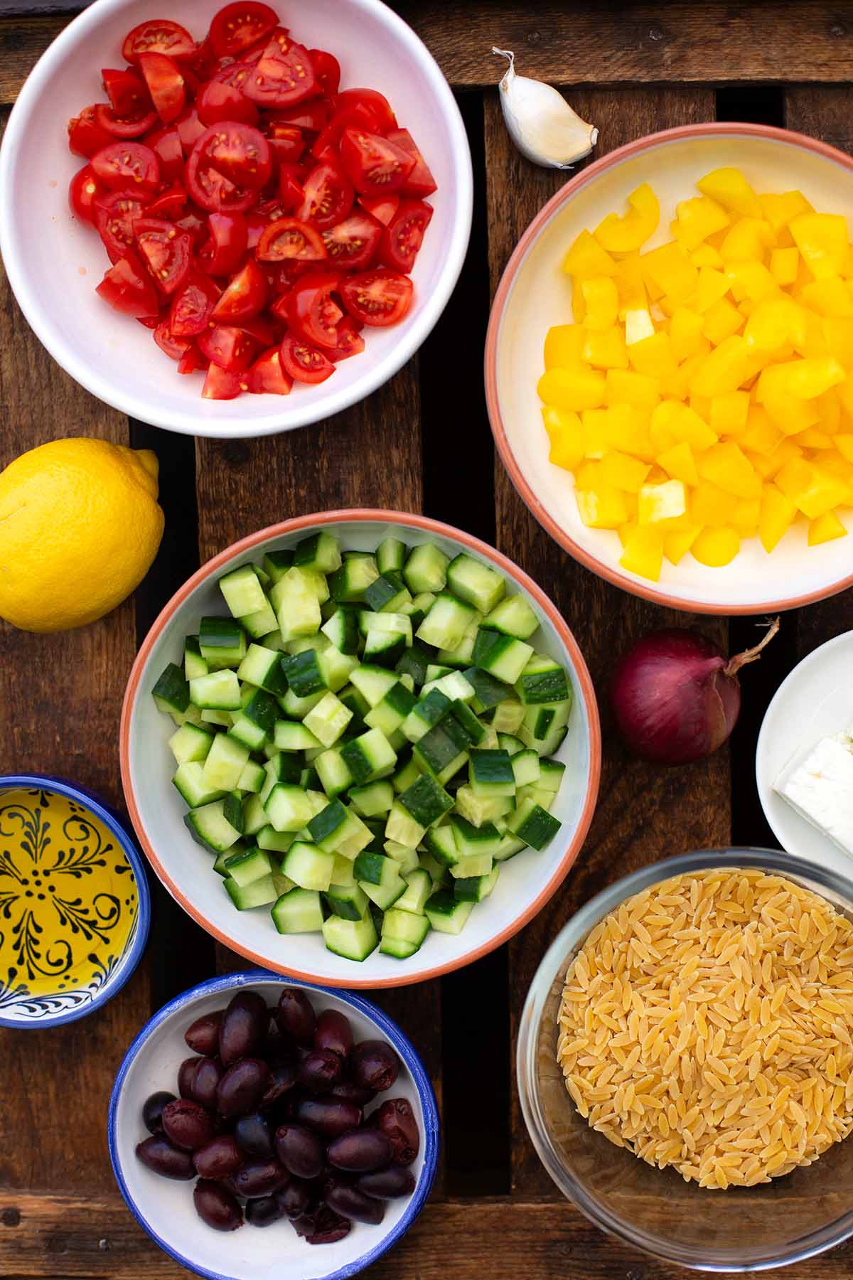 Zutaten in einzelnen kleinen Schüsseln für den griechischen Nudelsalat auf dunklem Holzuntergrund. Zu sehen sind gewürfelte Gurke, Tomaten und gelbe Paprika, eine ganze Zitrone, rote Zwiebel, Feta, Olivenöl und Kritharaki Nudeln