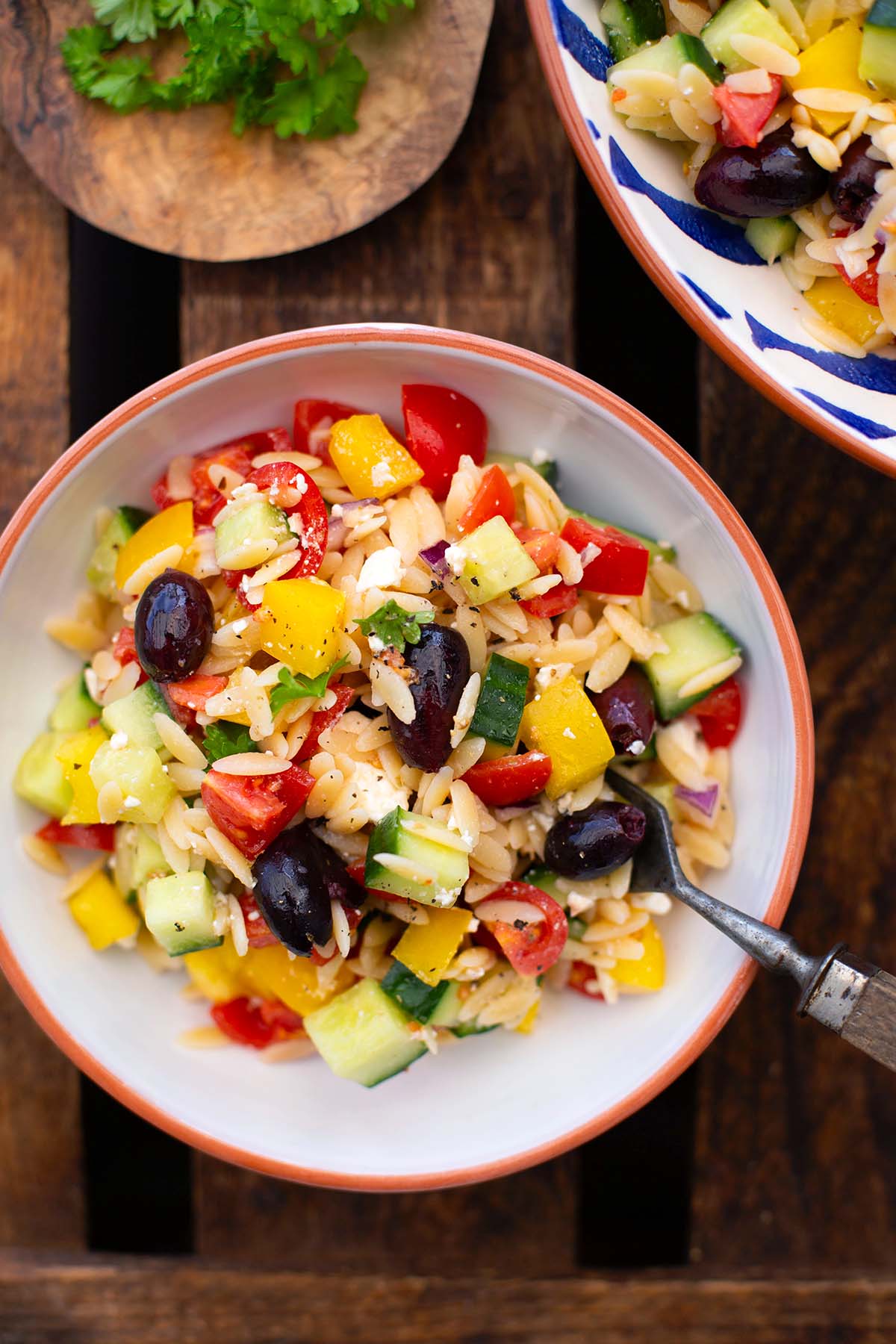 Fertiger Kritharaki Salat aufgeteilt auf eine große und eine kleine weiße Schüssel mit orangem Rand. Darin eine Gabel, bereit zum Essen. Daneben ein kleines Holzbrett mit Petersilie.