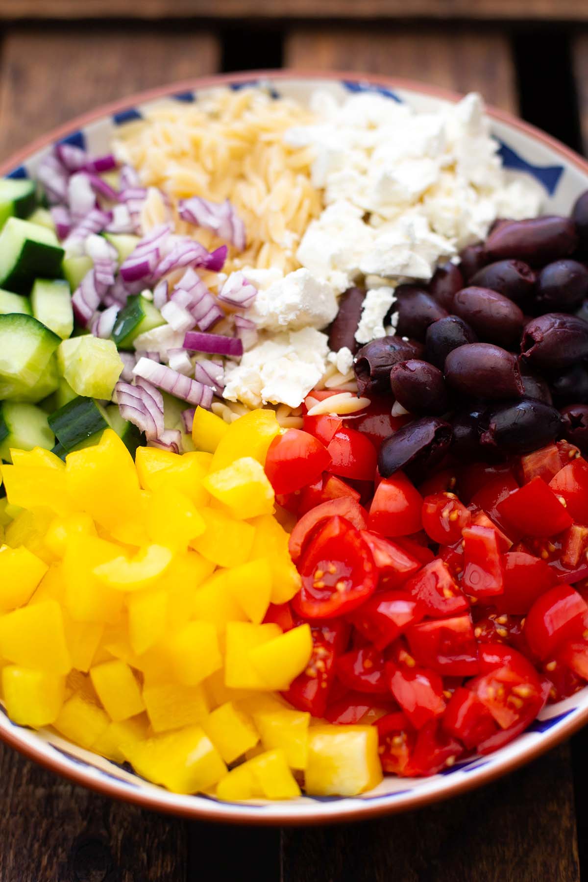 Gewürfelte Zutaten für den Kritharaki Salat in einer Schüssel. Zu sehen sind rote Zwiebel, Feta, Tomaten, gelbe Paprika, Gurke und schwarze Oliven.