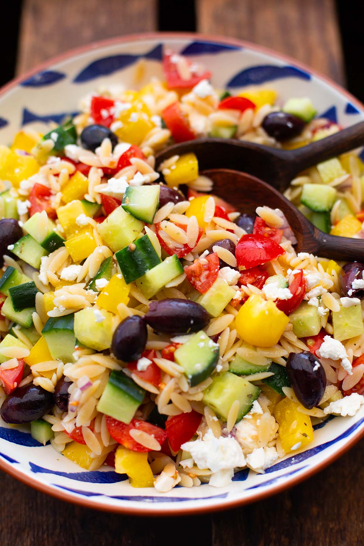 Fertiger Kritharaki Salat in einer weißen Schüssel mit braunem Rand und blauen Fischen. Die Schüssel steht auf dunklem Holzuntergrund. Zwei Holzlöffel zum Servieren.