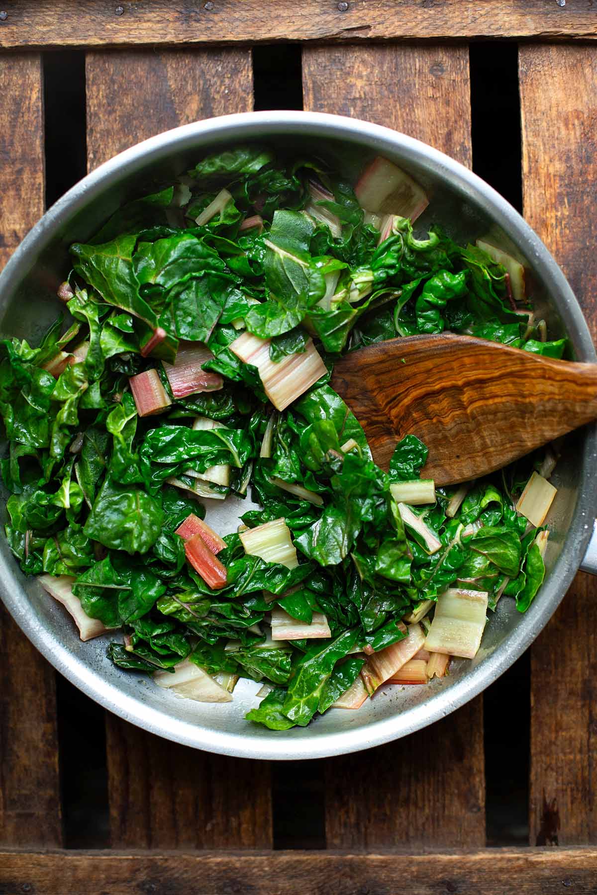 Klein geschnittener Mangold (Blätter und Stiele) in einer silbernen Pfanne auf dunklem Holzuntergrund. Dazu ein brauner Pfannenwender aus Holz.