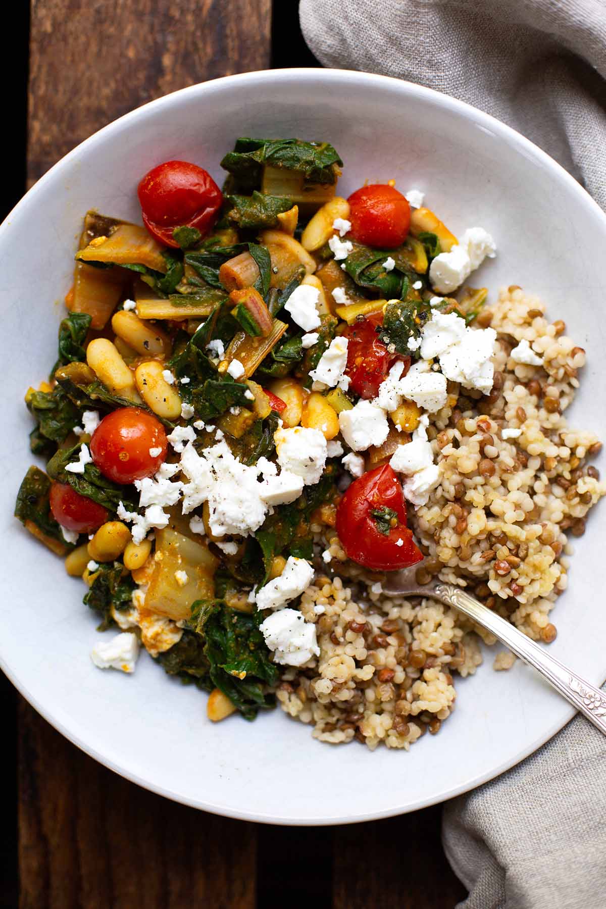Fertige Mangold-Pfanne mit Reis als Beilage, in einem weißen tiefen Teller. Garniert mit Feta-Krümeln. Der Teller, mit einer silbernen Gabel, steht auf einem dunklen Holzuntergrund.