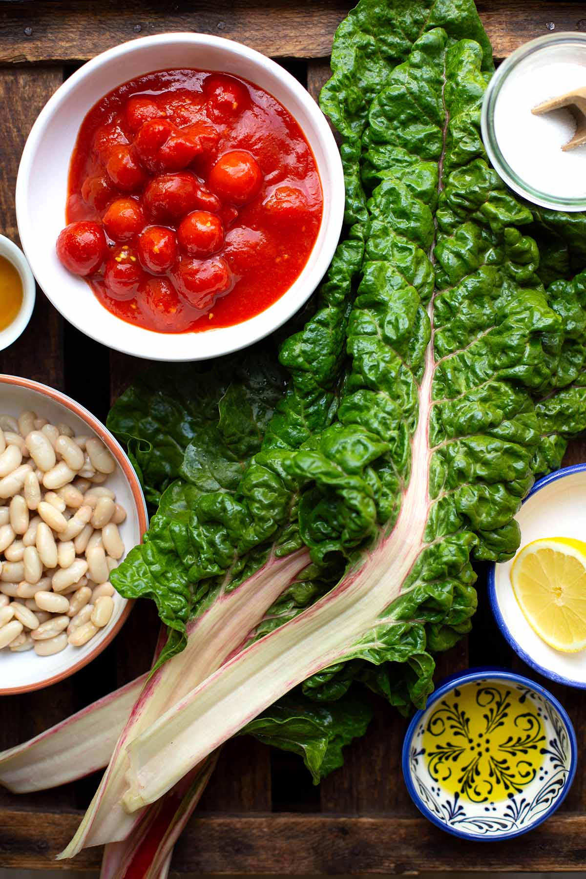 Zutaten für die einfache Mangold-Pfanne. Zu sehen sind Mangold, Olivenöl, weiße Bohnen, eine halbe Zitrone, Cherrytomaten aus der Dose, Ahornsirup und Salz.