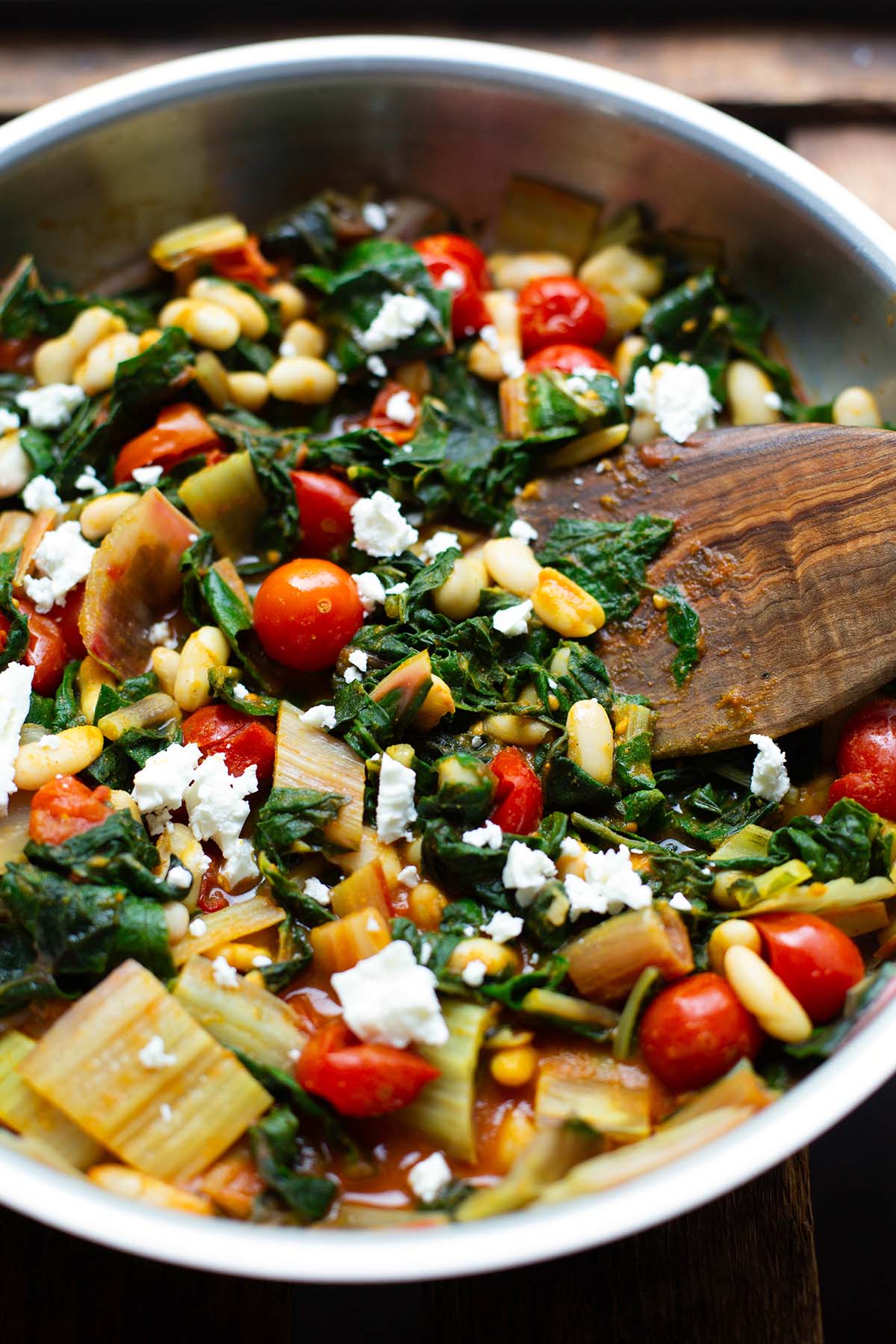 Fertige Mangold-Pfanne in einer silbernen Pfanne, garniert mit Feta-Krümeln. Die Pfanne steht auf einem dunklen Holzuntergrund. In der Pfanne liegt ein Pfannenwender aus Holz.