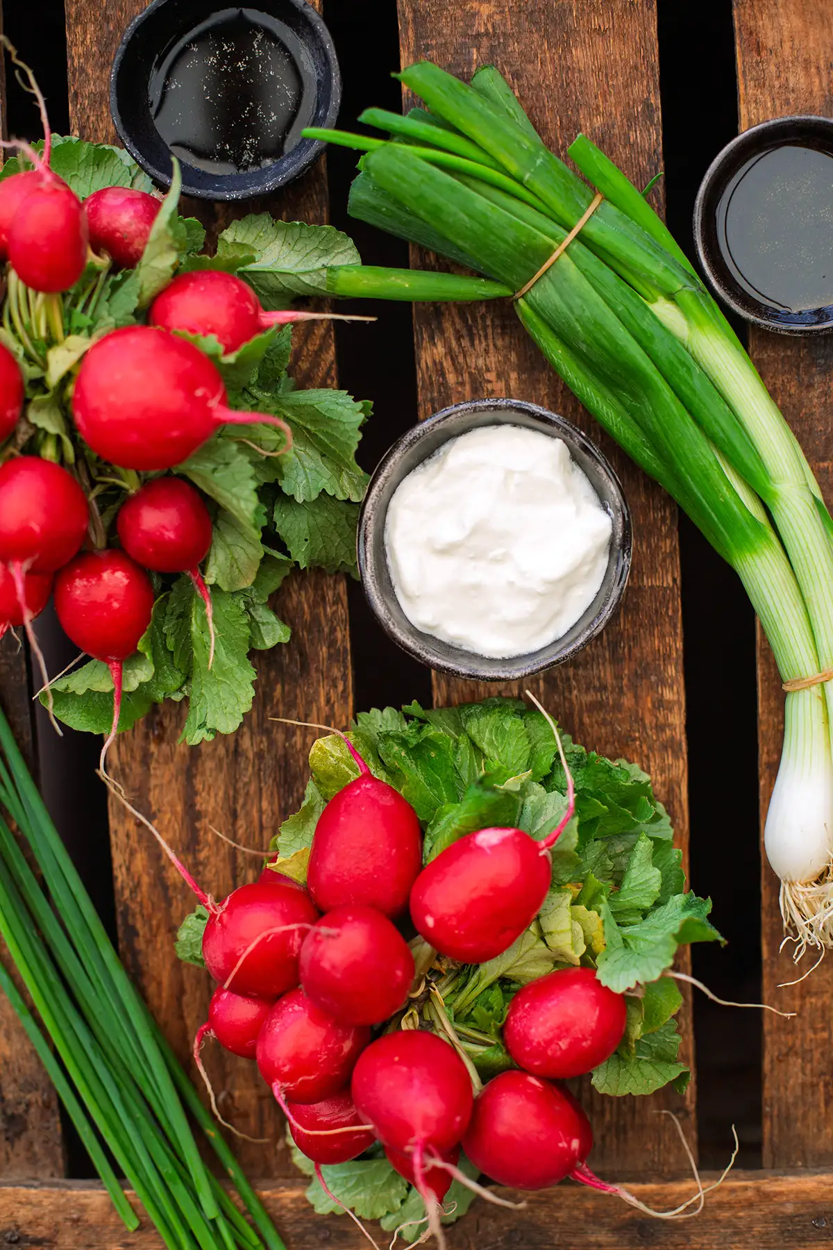 Einzelne Zutaten für den Radieschensalat wie von Oma auf dunklem Holzuntergrund. Zu sehen sind zwei Bund Radieschen, Frühlingszwiebeln, saure Sahne, Schnittlauch, Essig und Öl.