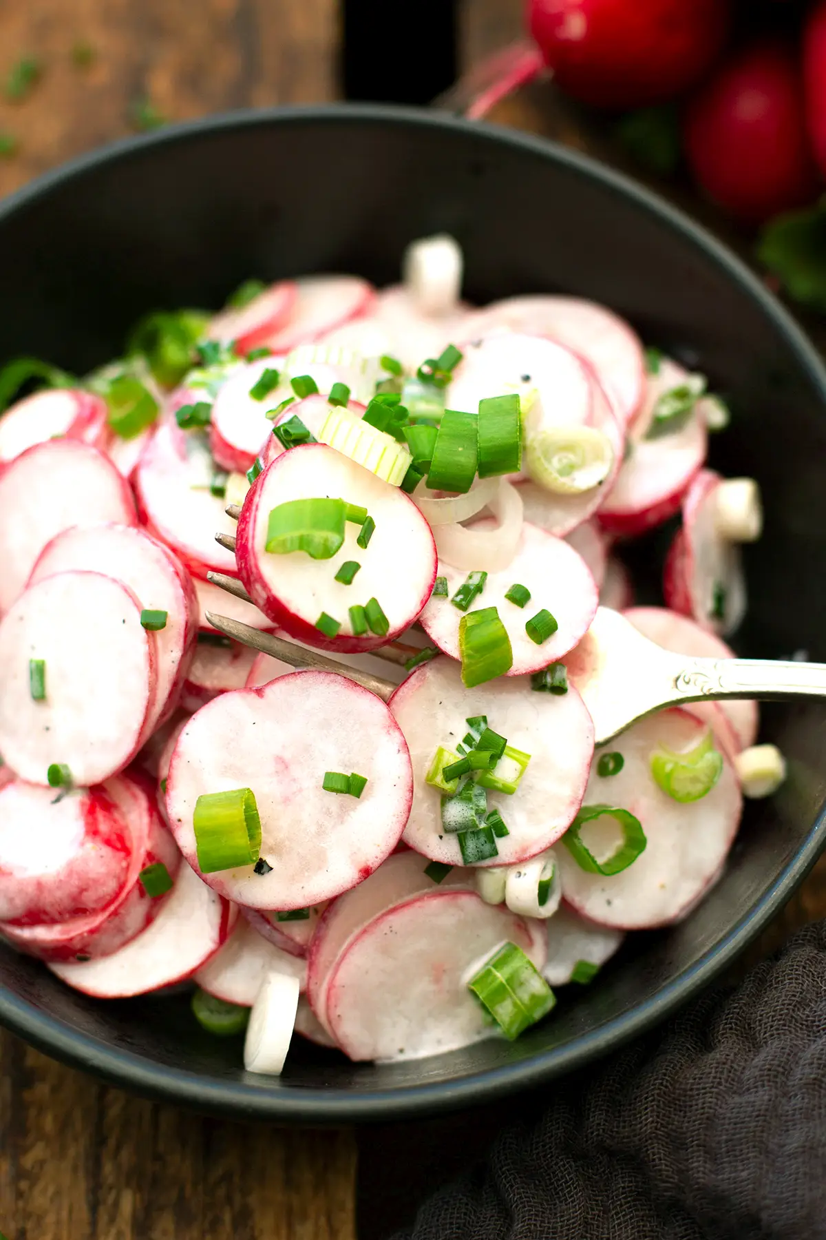Omas Radieschensalat in einer schwarzen Schüssel zusammen mit einer Gabel. Die Schüssel steht auf einem dunklen Holzuntergrund.