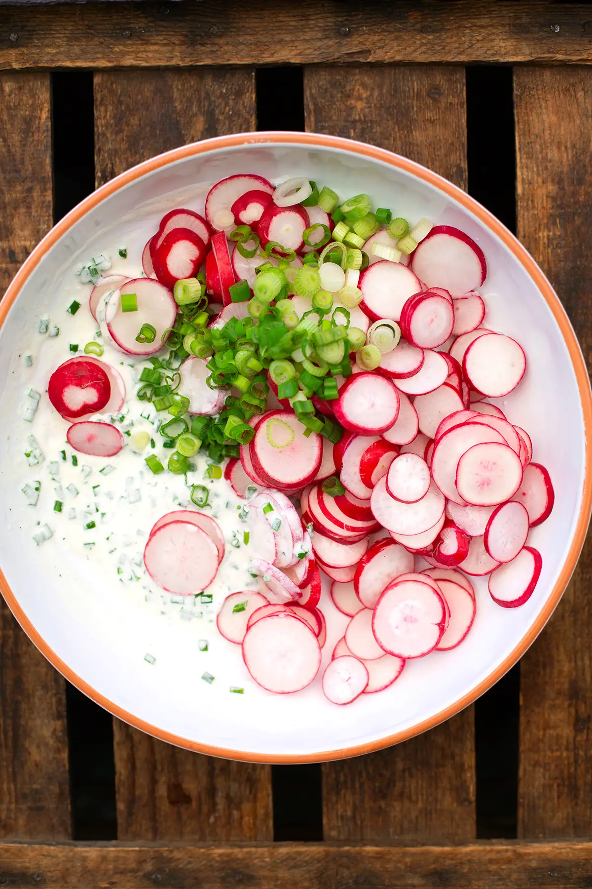 In Scheiben geschnittene Radieschen, Frühlingszwiebeln, Schnittlauch und das fertige Dressing für den Radieschensalat mit Sahne in einer weißen Schüssel mit orangem Rand. Die Schüssel steht auf einem dunklen Holzuntergrund.