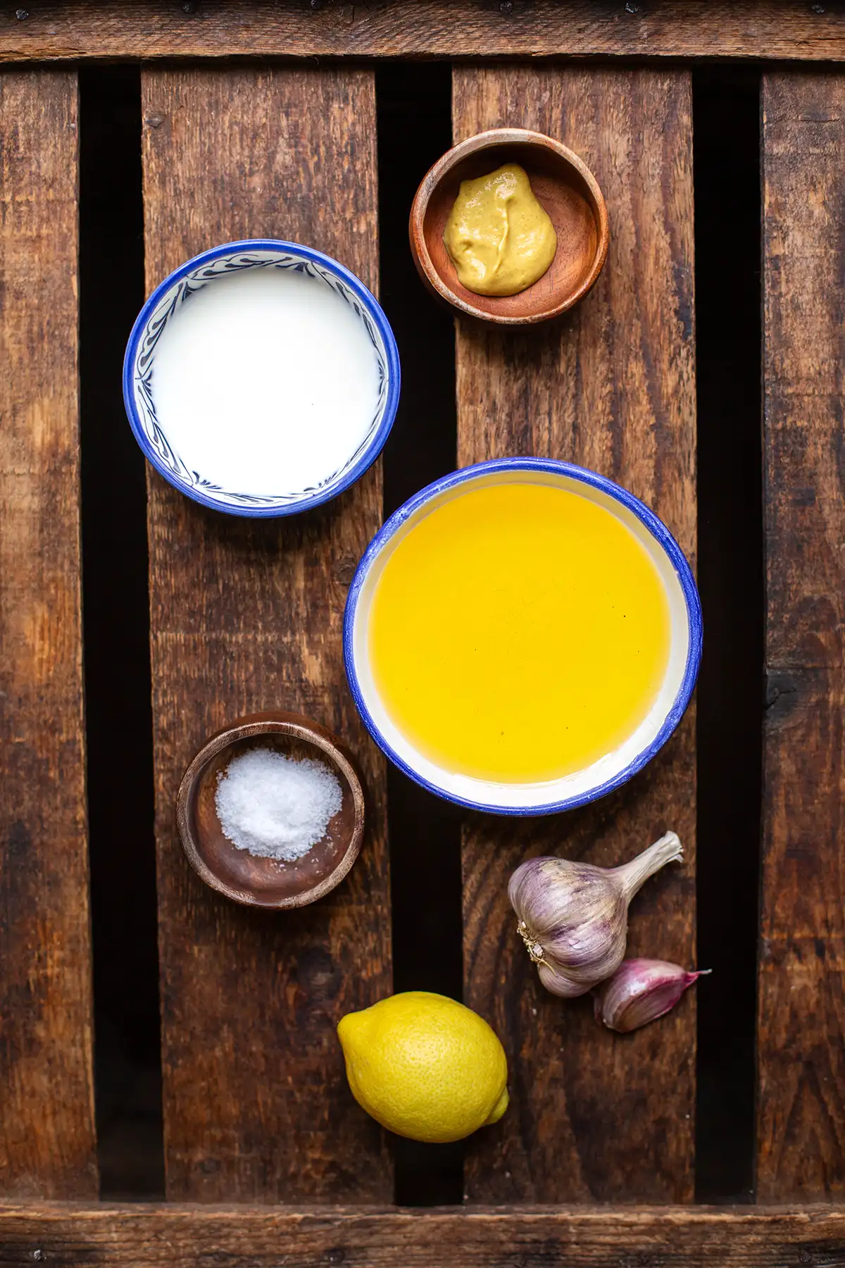 Die Zutaten für selbstgemachte Aioli ohne Ei einzeln auf Schüsseln aufgeteilt. Zu sehen sind Olivenöl, Knoblauchzehen, Salz, Zitrone, Milch und Senf.