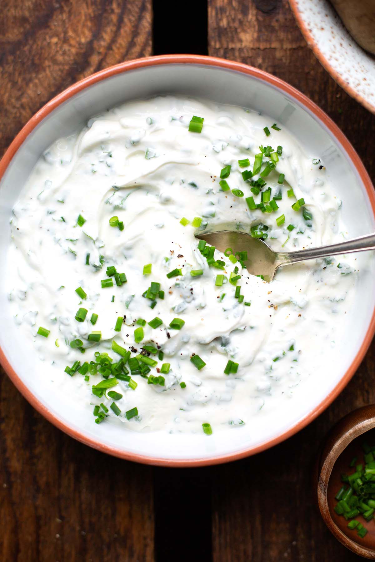 Fertiger Kräuterquark in einer weißen Schüssel mit orangem Rand. Garniert mit gehacktem Schnittlauch und mit einem silbernen Löffel. Die weiße Schüssel steht auf einem dunklen Holzuntergrund.
