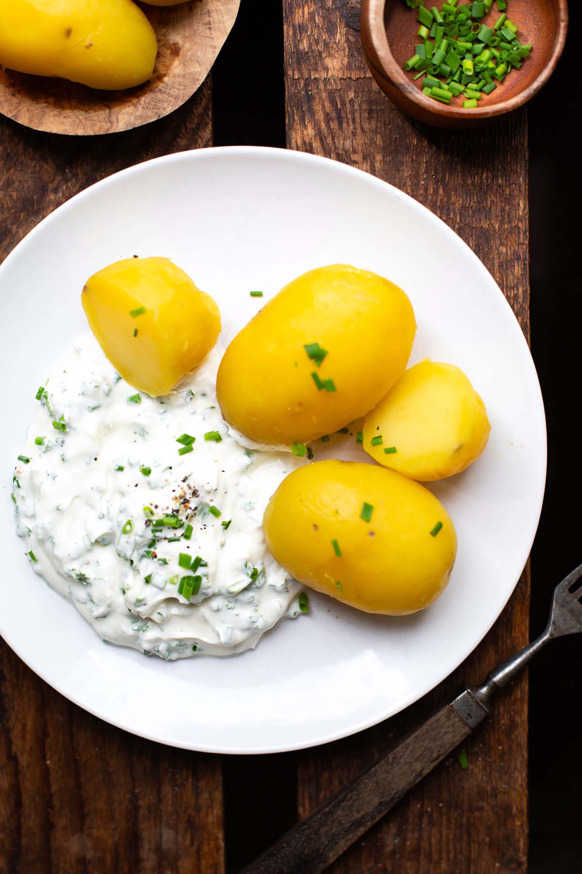Fertige Kartoffeln mit Quark auf einem weißen Teller, der auf einem dunklen Holzuntergrund steht. Neben dem Teller liegt eine Gabel und zwei Schüsseln mit einmal geschälten Kartoffeln und einmal gehackte Petersilie.