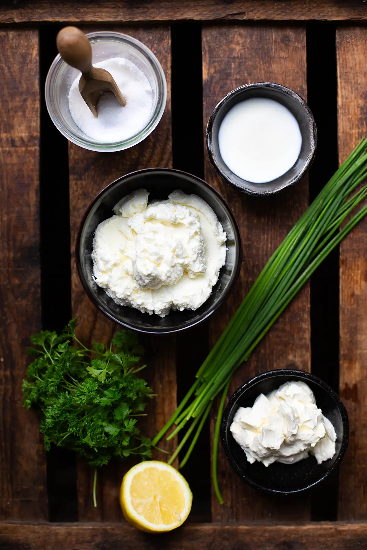 Zutaten für cremigen Kräuterquark einzeln in Schüsselchen oder lose auf einem dunklen Holzuntergrund. Zu sehen sind, Schmand, Speisequark, Schnittlauch, Petersilie, eine halbe Zitrone, Milch und Salz.