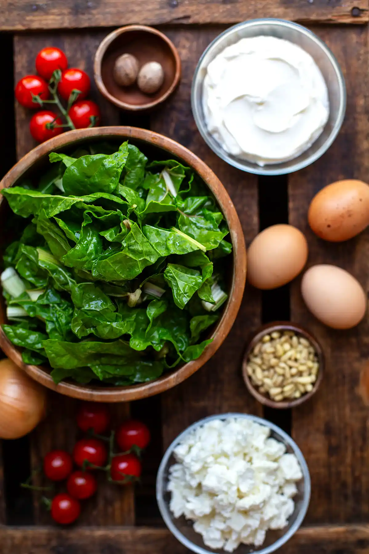 Zutaten für die vegetarische Mangold-Quiche einzeln in Schüsseln oder lose auf einem dunklen Holzuntergrund. Zu sehen sind klein geschnittener Mangold, drei Eier, Schmand, Pinienkerne, Cherrytomaten, Muskatnuss, Fetakrümel und eine weiße Zwiebel.